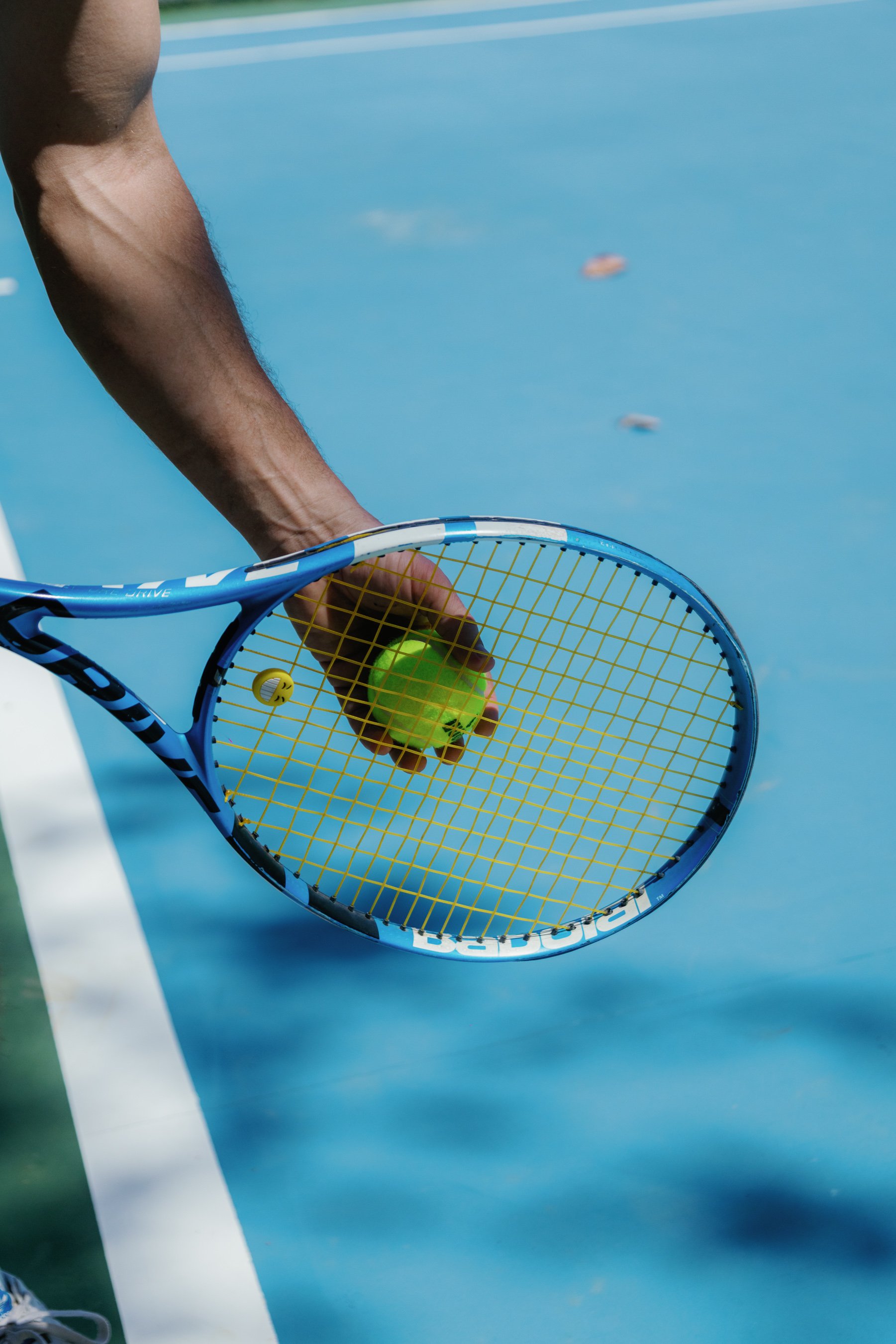 Close-up of a person holding a green tennis ball with a tennis racket on a blue tennis court.