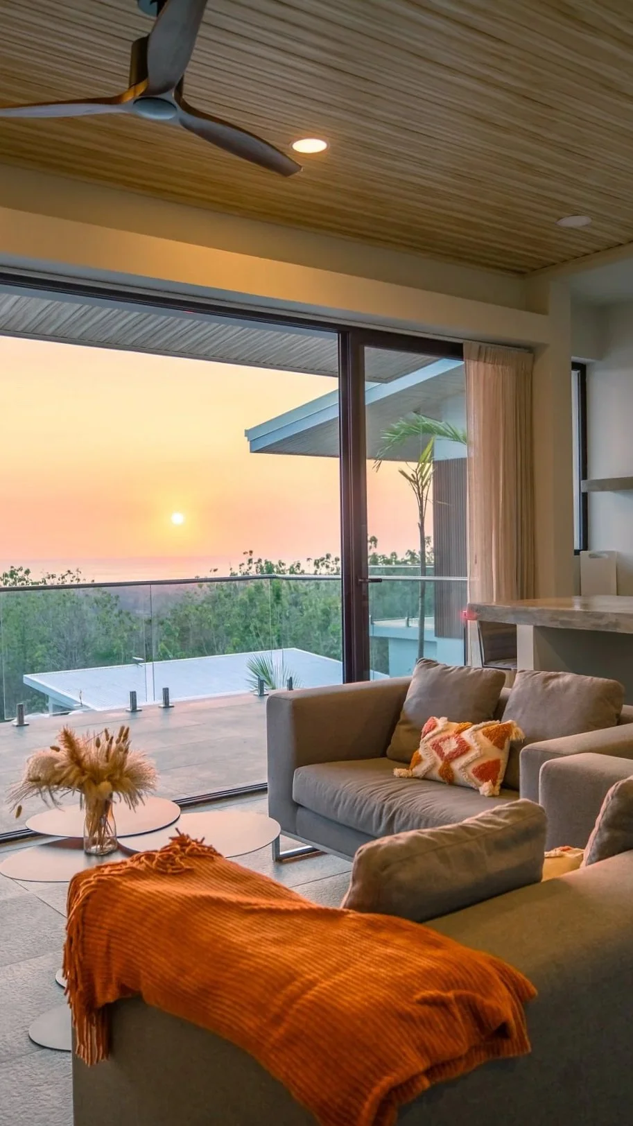 Living room with large glass sliding door opening to a balcony with a sunset view of trees and a calm ocean. Indoor decor includes a beige sofa with decorative pillows, a coffee table with a vase of dried flowers, and a cozy orange throw blanket. The ceiling features wood paneling and a ceiling fan.