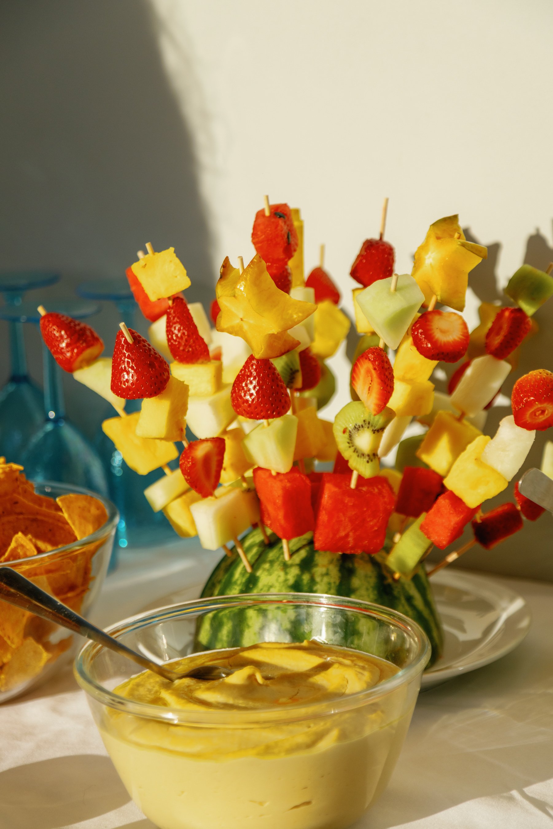Fruit skewers with strawberries, pineapple, cantaloupe, watermelon, honeydew, kiwi, and starfruit arranged in a watermelon bowl, with a bowl of yellow dip in front.