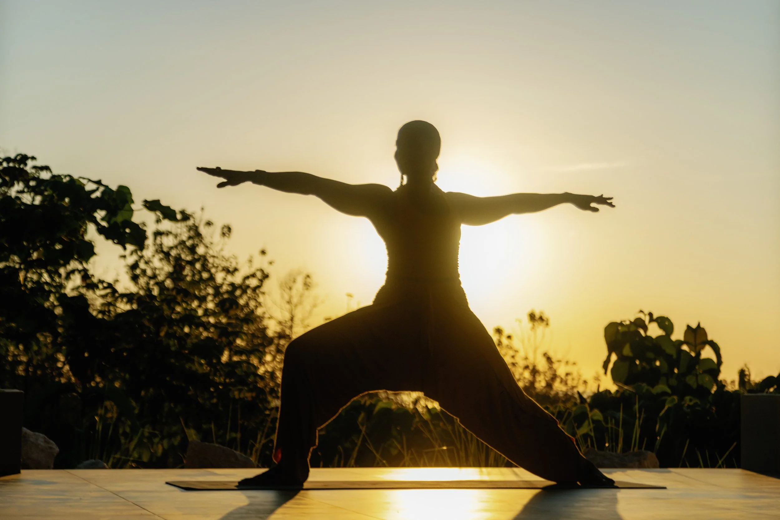 Silhouette of a person practicing yoga outdoors at sunset, standing in a Warrior pose with arms extended and one leg bent, surrounded by trees and plants.