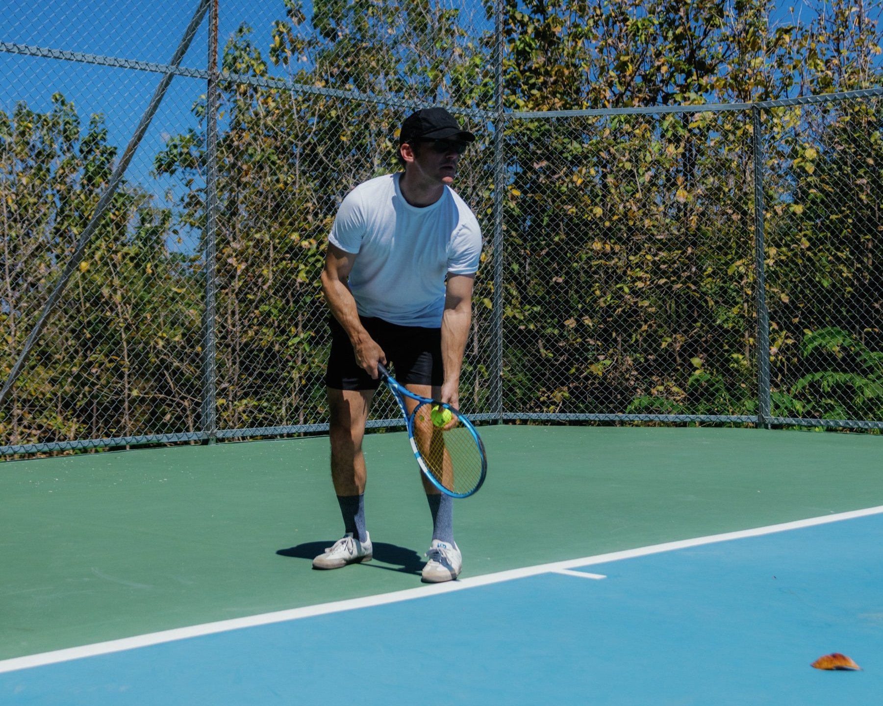Person playing tennis on an outdoor court, holding a tennis racket and ready to hit a tennis ball, with trees and a chain-link fence in the background.