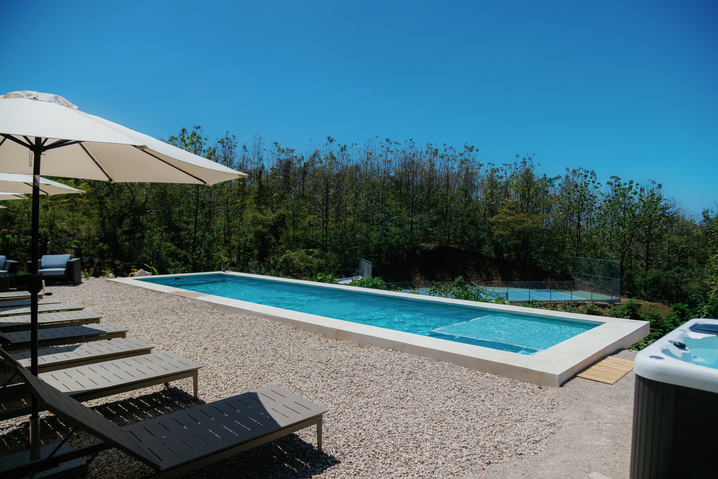 Poolside area with lounge chairs, umbrellas, and a rectangular swimming pool, surrounded by gravel and trees under a clear blue sky.