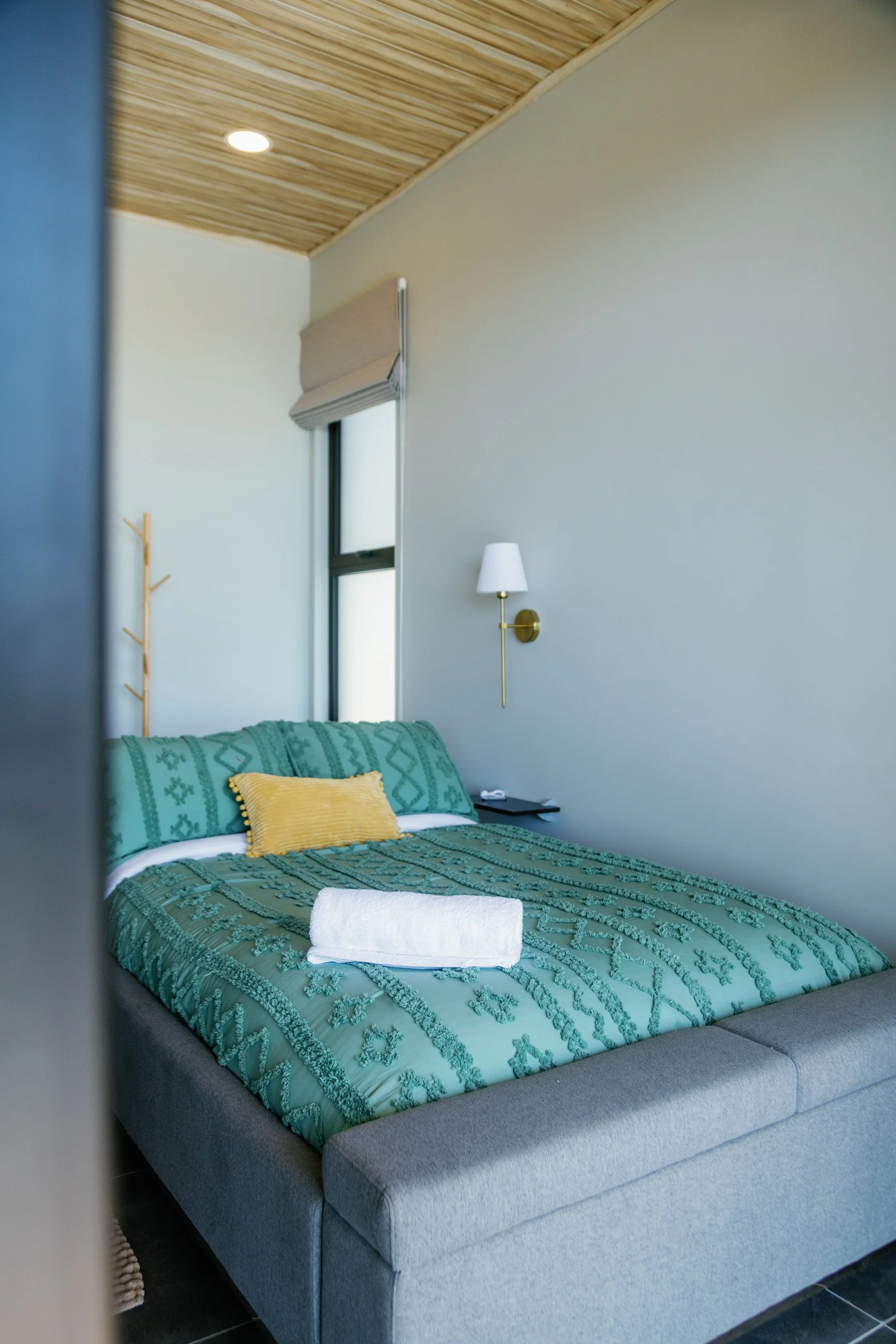 A neatly made bed with a green patterned quilt, a yellow pillow, and a rolled white towel on top. The room has a window with a beige Roman shade, a white wall, a wall-mounted white lamp with a brass arm, a small black bedside table with a telephone, 