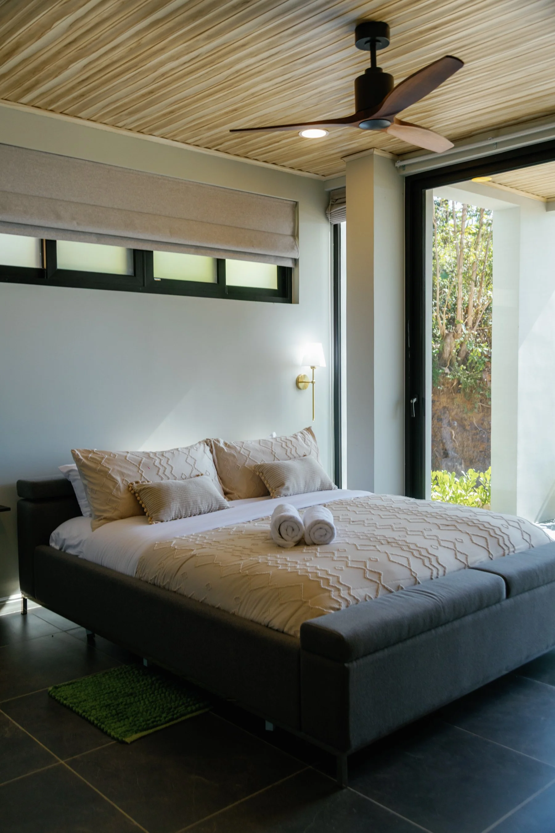 Modern bedroom with a dark gray bed, beige bedding, two rolled towels, a green rug, a wall-mounted light, and a large window showing outdoor trees.