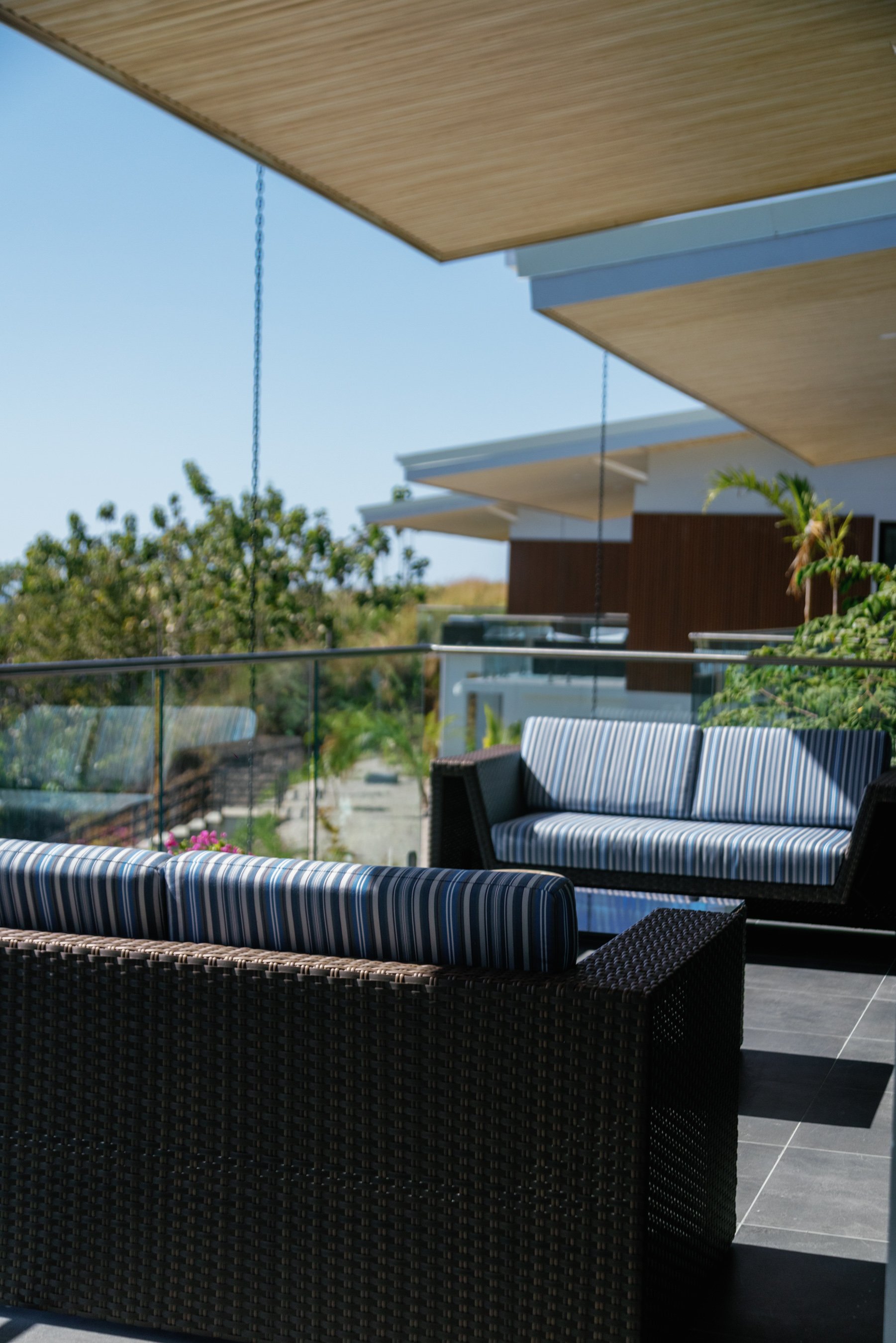 A modern outdoor patio with black rattan furniture and striped cushions, under large beige umbrellas, overlooking a garden with green plants and trees.