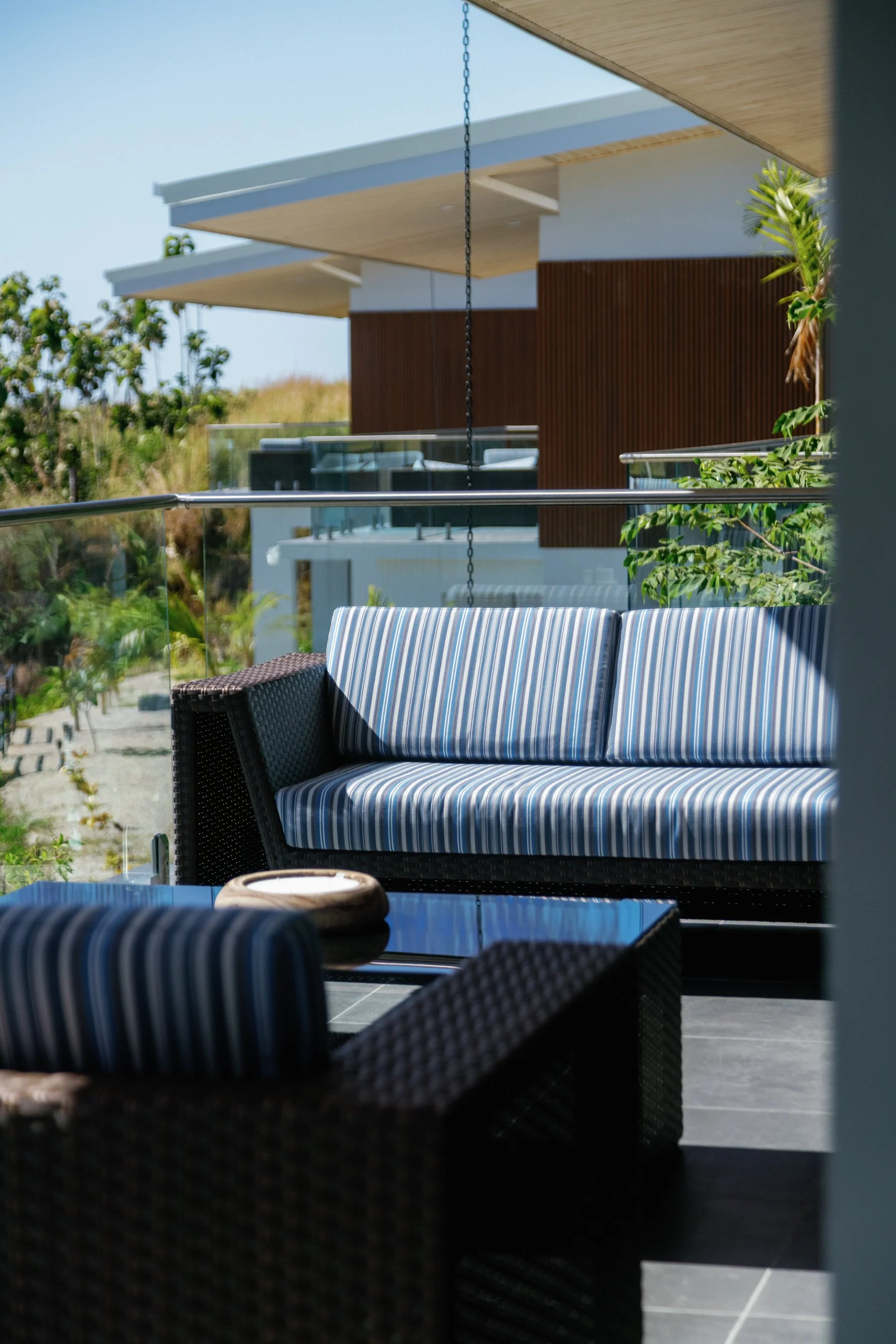 Outdoor patio area with rattan furniture, including a striped cushioned sofa and a matching chair, under a covered area with modern architecture, glass railing, and greenery in the background.