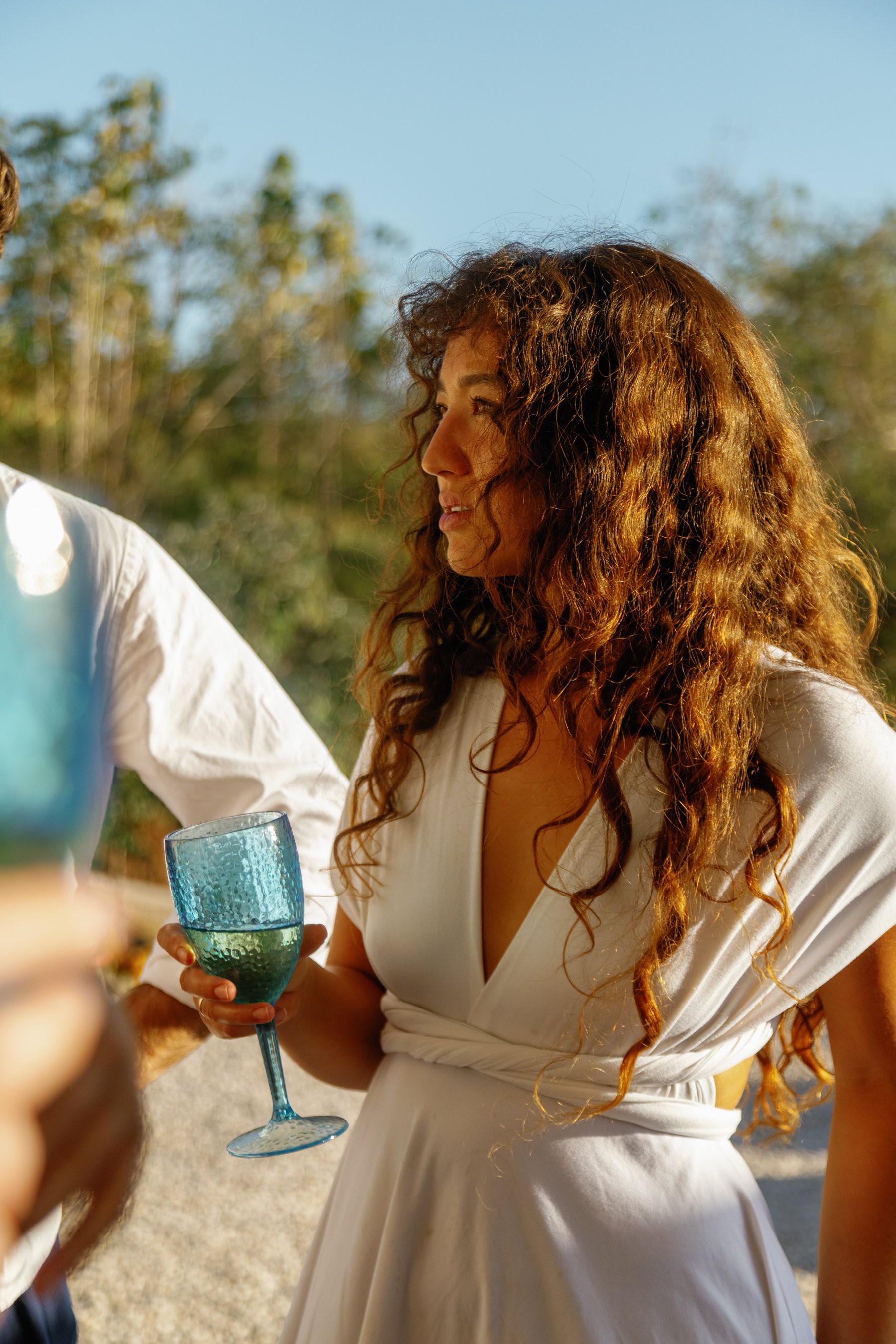 A woman with long, curly hair holds a blue glass of wine at an outdoor gathering.