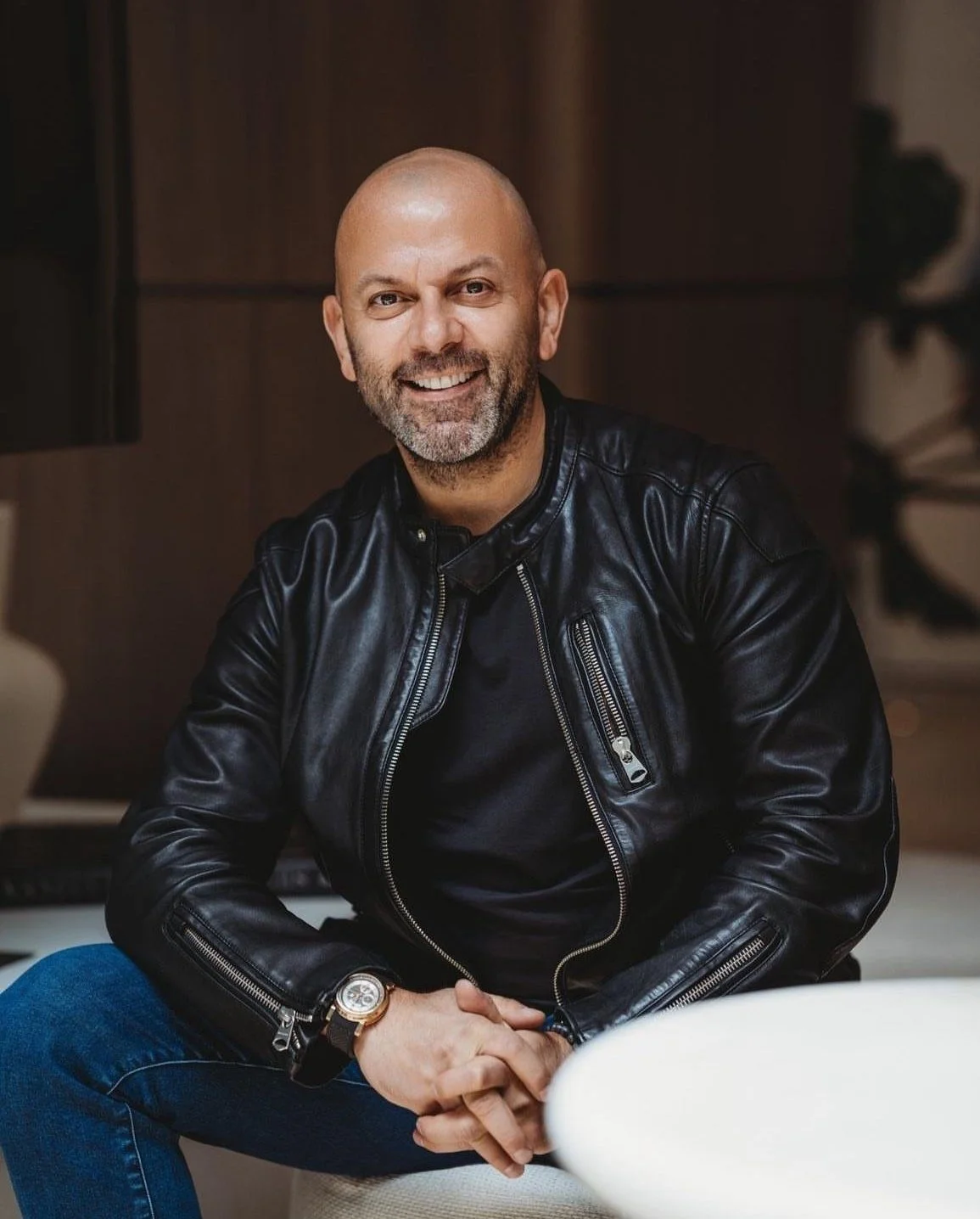 A smiling middle-aged man with a beard, wearing a black leather jacket and a wristwatch, sitting indoors.