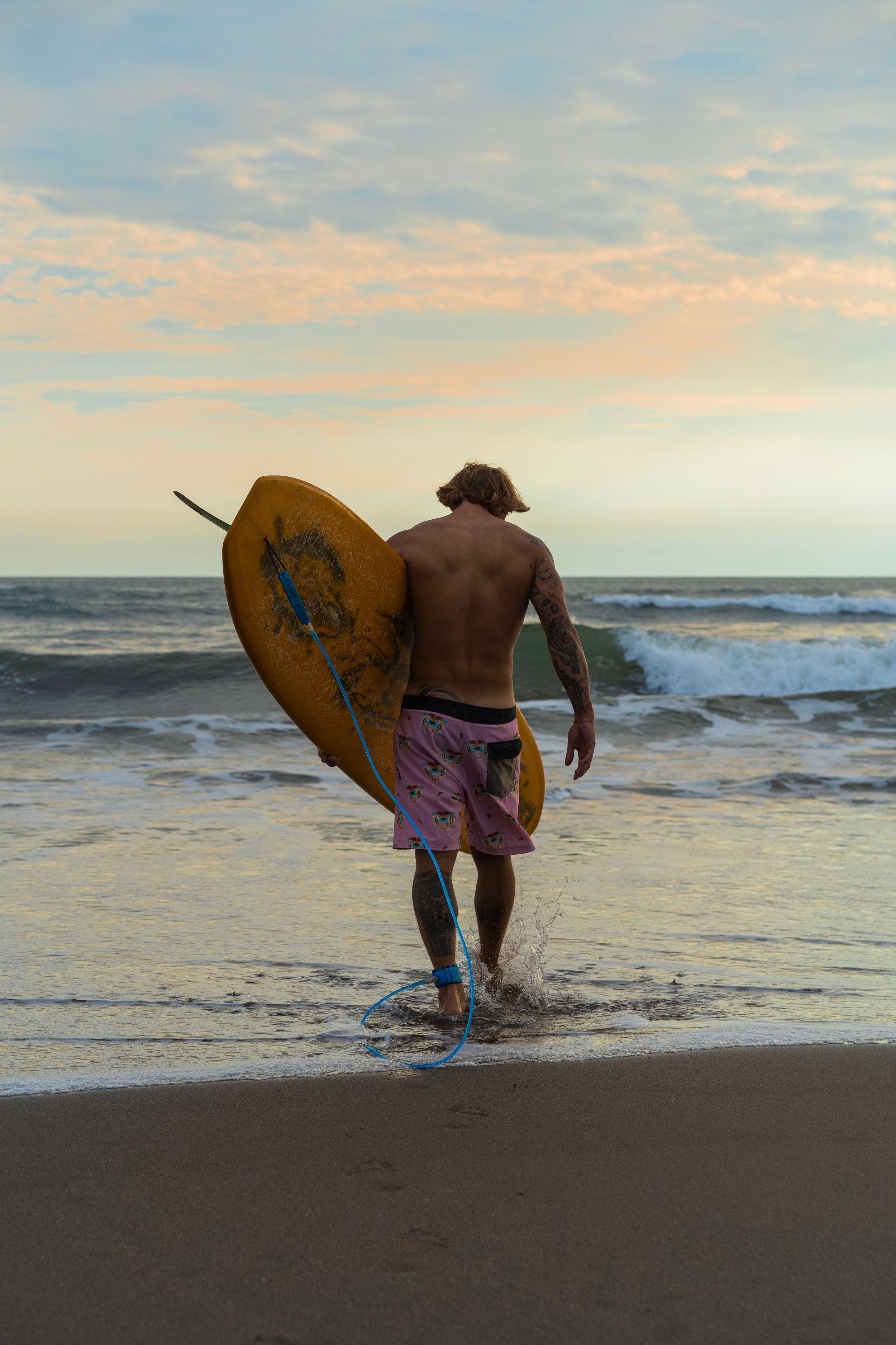 A man with tattoos walking into the ocean holding a yellow bodyboard with a leash in pink and blue, at sunset or sunrise.