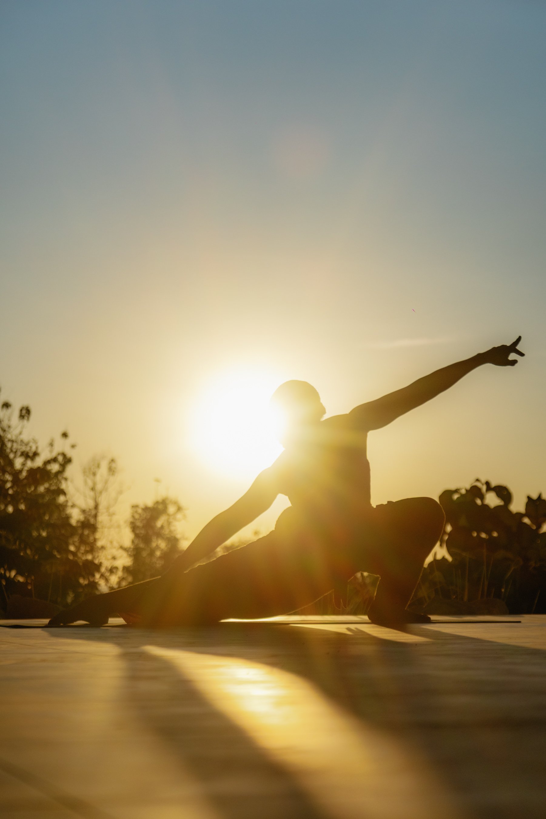 A person doing a yoga stretch outdoors during sunset, sitting on the ground with one leg extended and the other bent, reaching one arm forward in a lunge pose, with the sun setting behind them.
