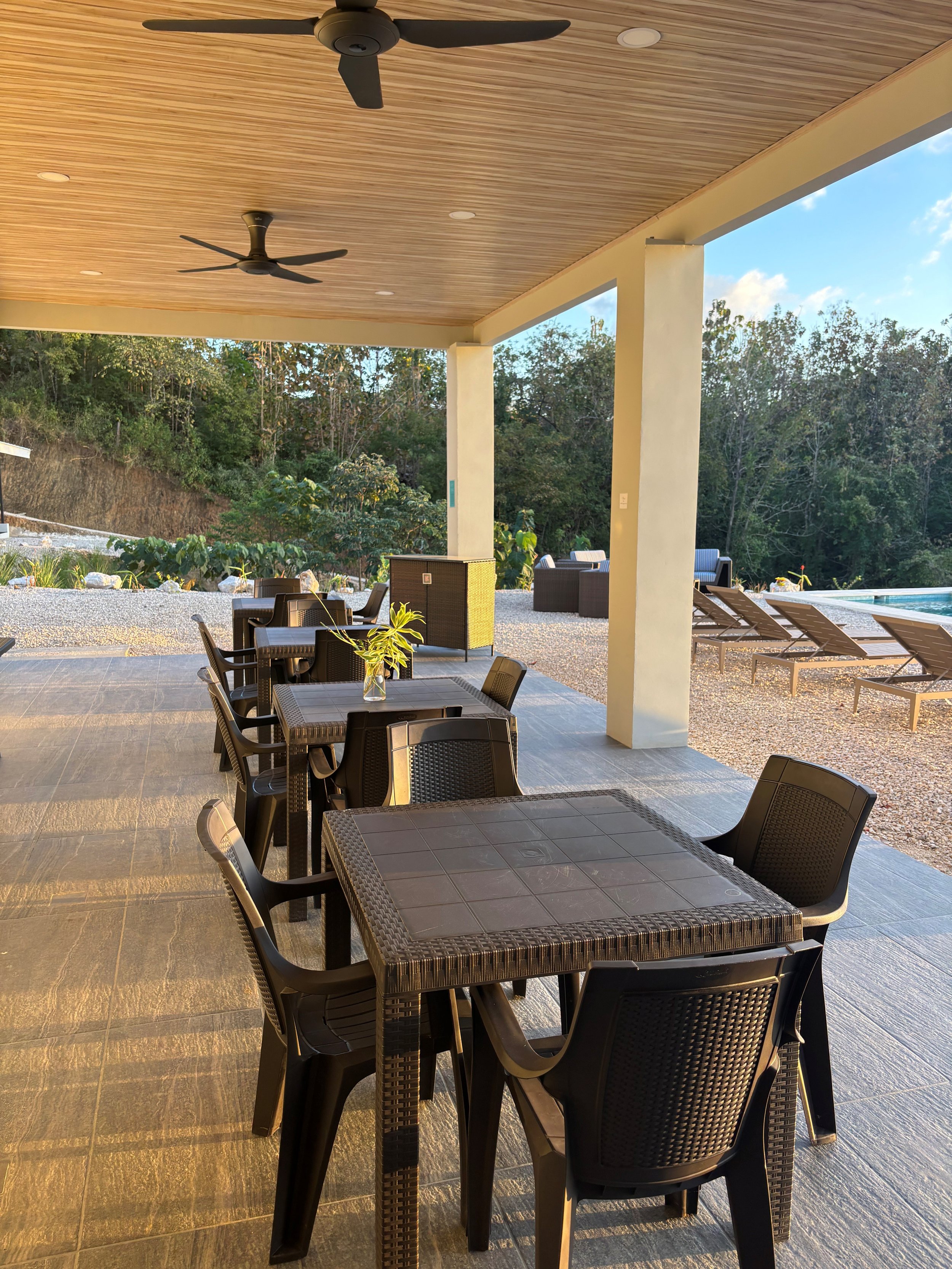 Outdoor patio with black tables and chairs, a potted plant, poolside lounge chairs, and lush greenery in the background.