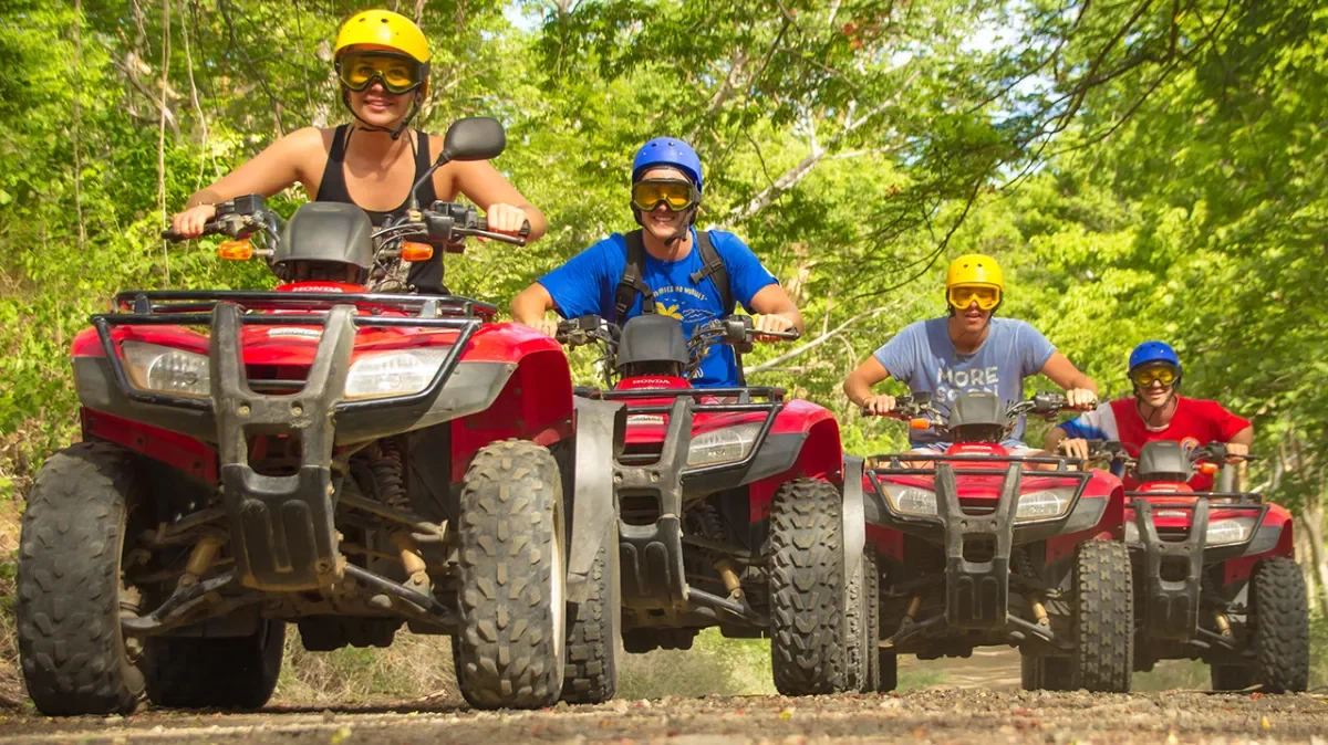 Four people riding all-terrain vehicles (ATVs) on a forest trail, wearing helmets and goggles, surrounded by green trees.