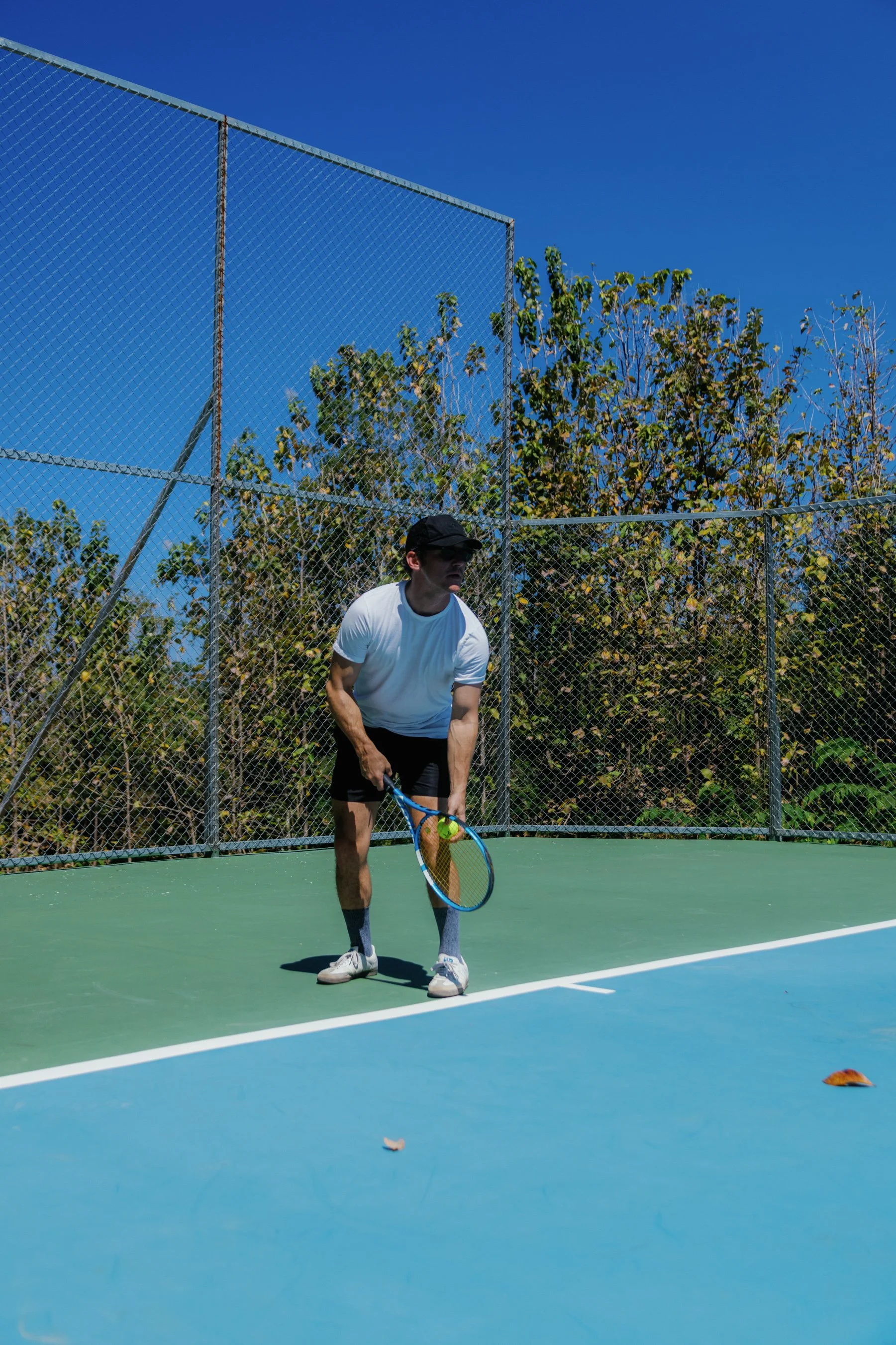 A man wearing a white shirt, black shorts, gray socks, white shoes, and a black cap, holding a tennis racket and preparing to hit a tennis ball on an outdoor court.