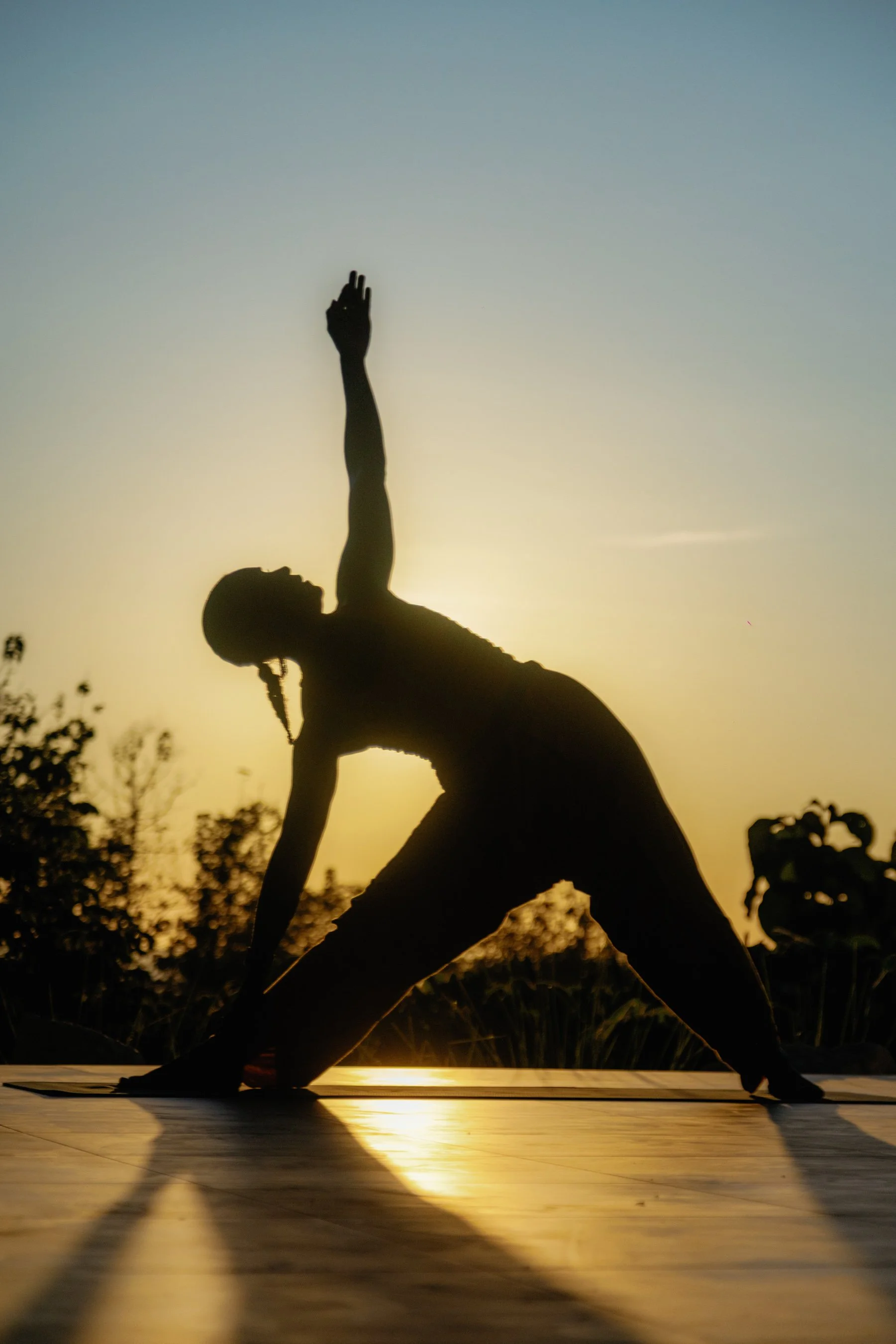 Silhouette of a woman practicing yoga on a mat during sunset, with one arm extended upward and the other hand on the ground.