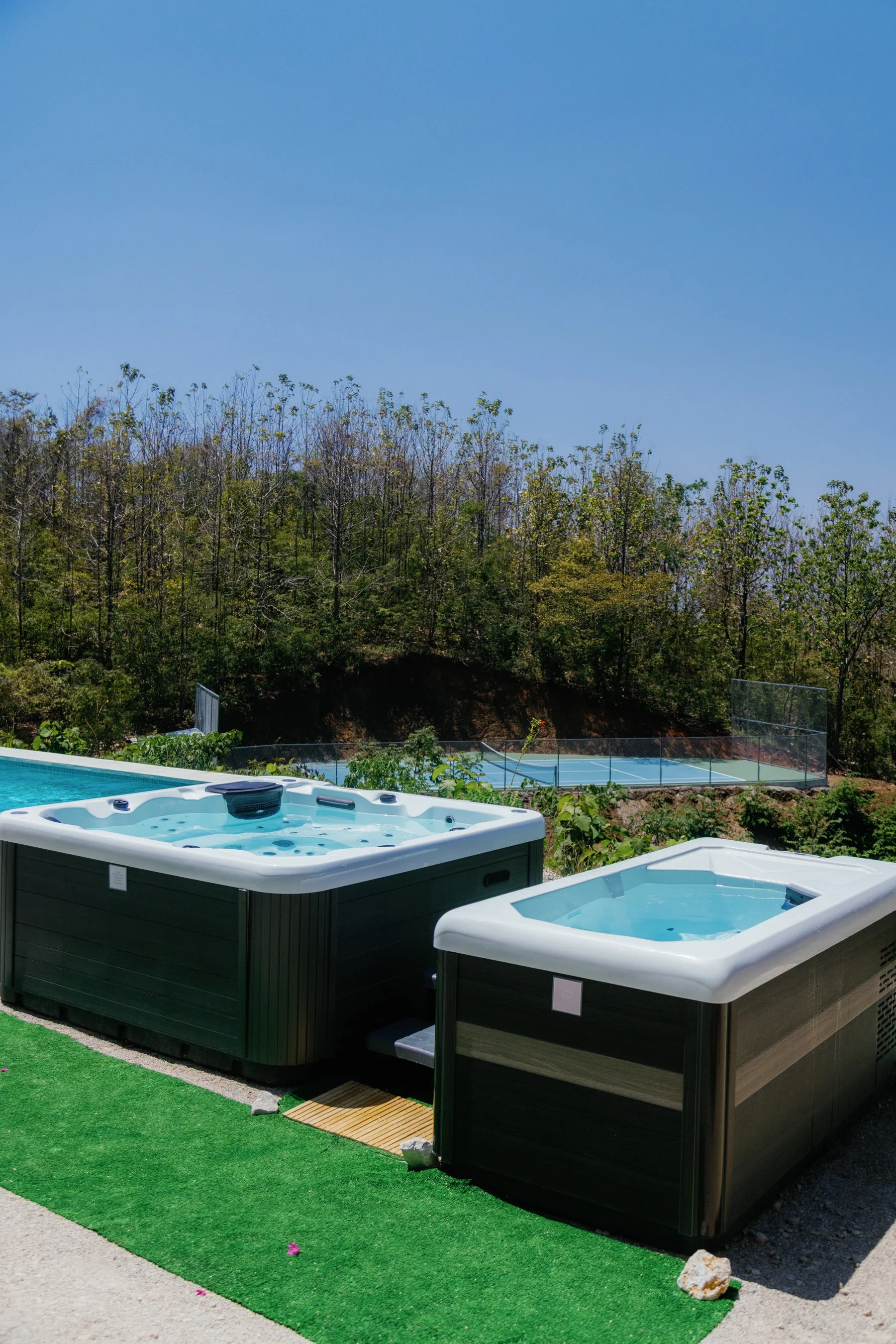 Two hot tubs on a patio with a swimming pool, tennis court, and wooded hillside in the background.