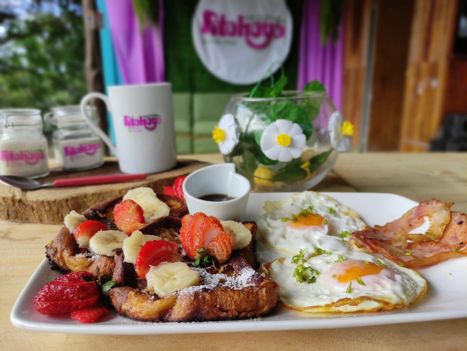 Plate with French toast topped with strawberries, bananas, and powdered sugar, sunny side up eggs, and a strip of bacon, served with syrup in a small container. In the background, there are coffee mugs, jars, and a bowl of flowers on a wooden table.