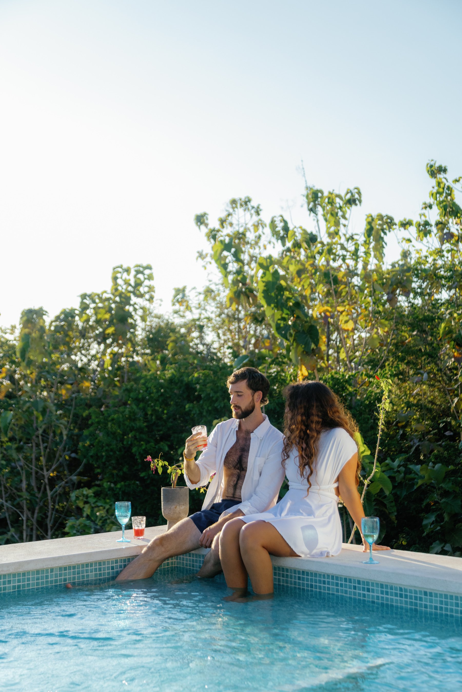A man and woman sit on the edge of a swimming pool with their feet in the water. The man is holding a drink, and there are glasses of beverages on the poolside. They are dressed casually, with the man in shorts and a partly unbuttoned shirt, and the 