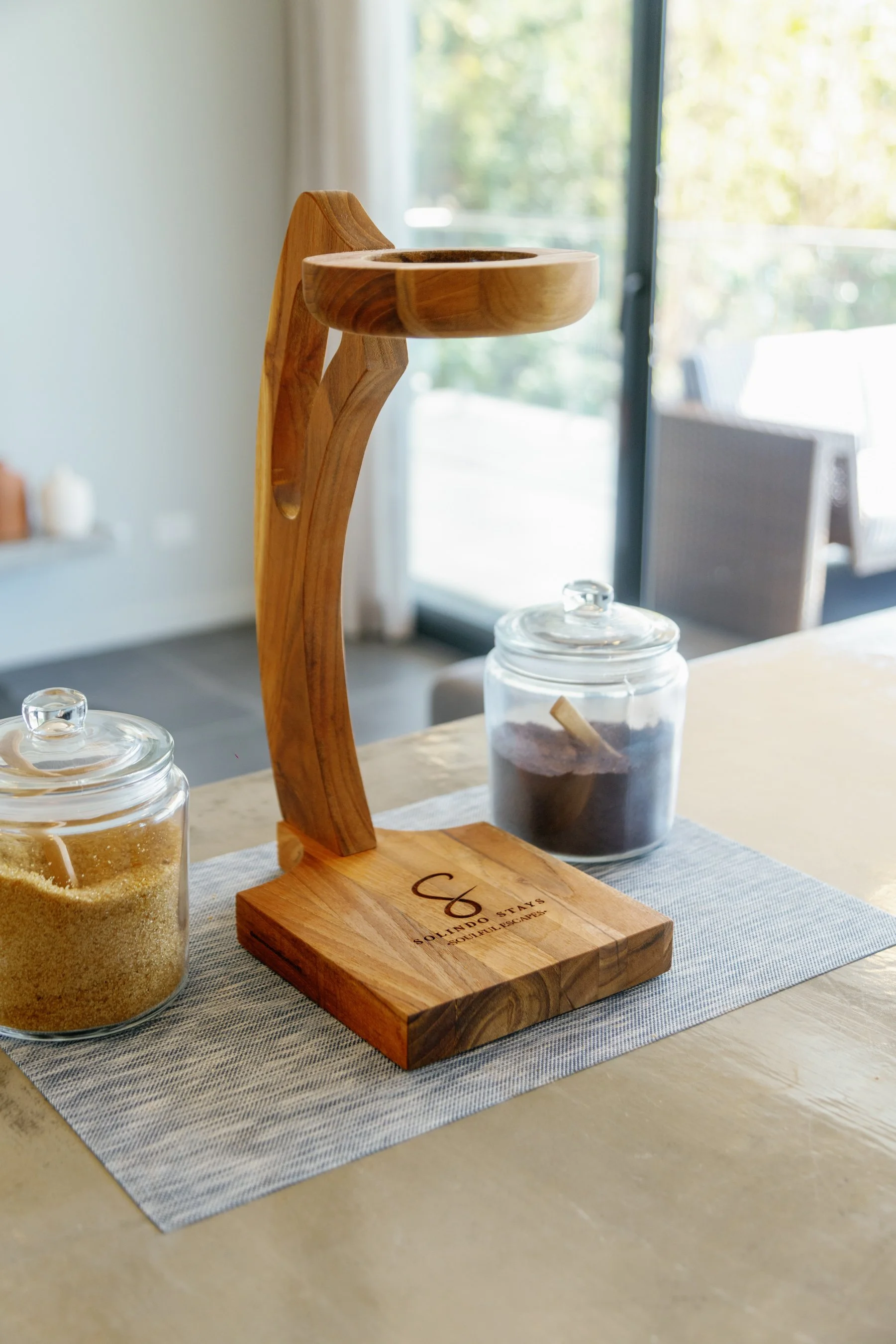 Decorative wooden stand with a circular tray on top, placed on a table with glass jars filled with sugar and coffee grounds, in a bright room with large windows.