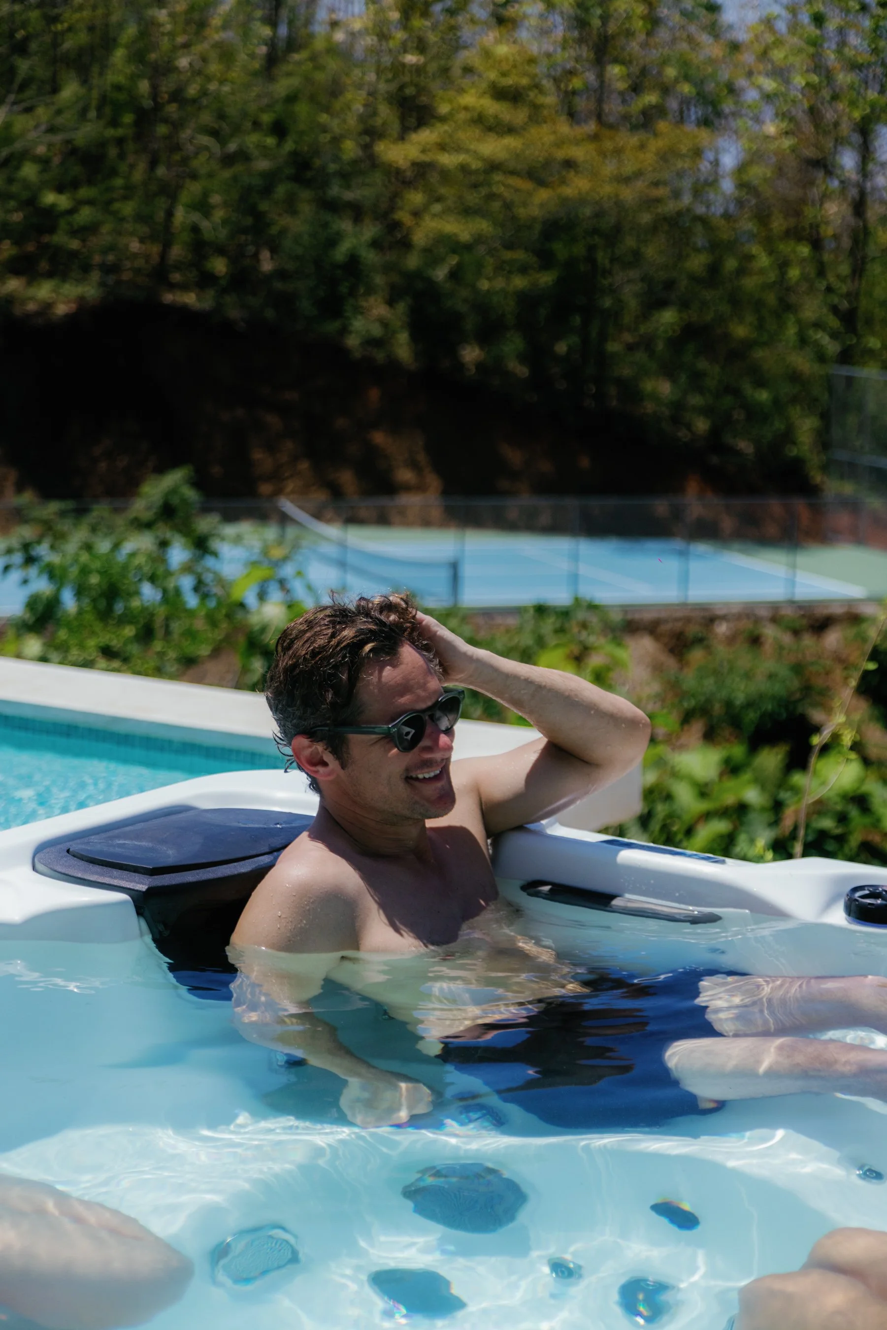 A man relaxing in a hot tub with sunglasses, smiling, and scratching his head on a sunny day with a tennis court and lush green trees in the background.