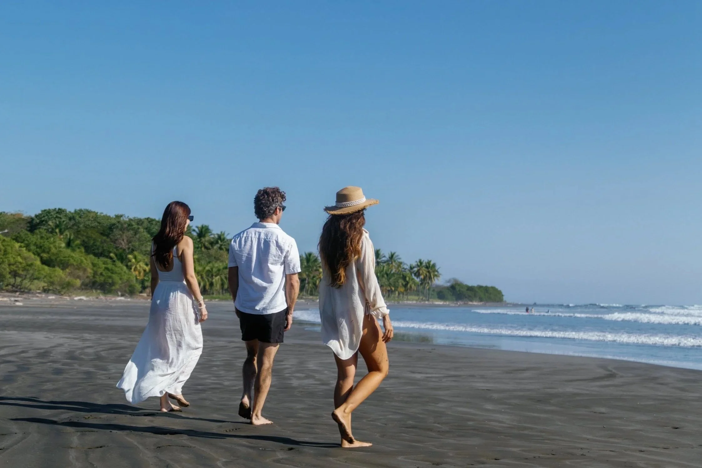 Three people walking on a beach, two women and one man, with ocean waves and green trees in the background.
