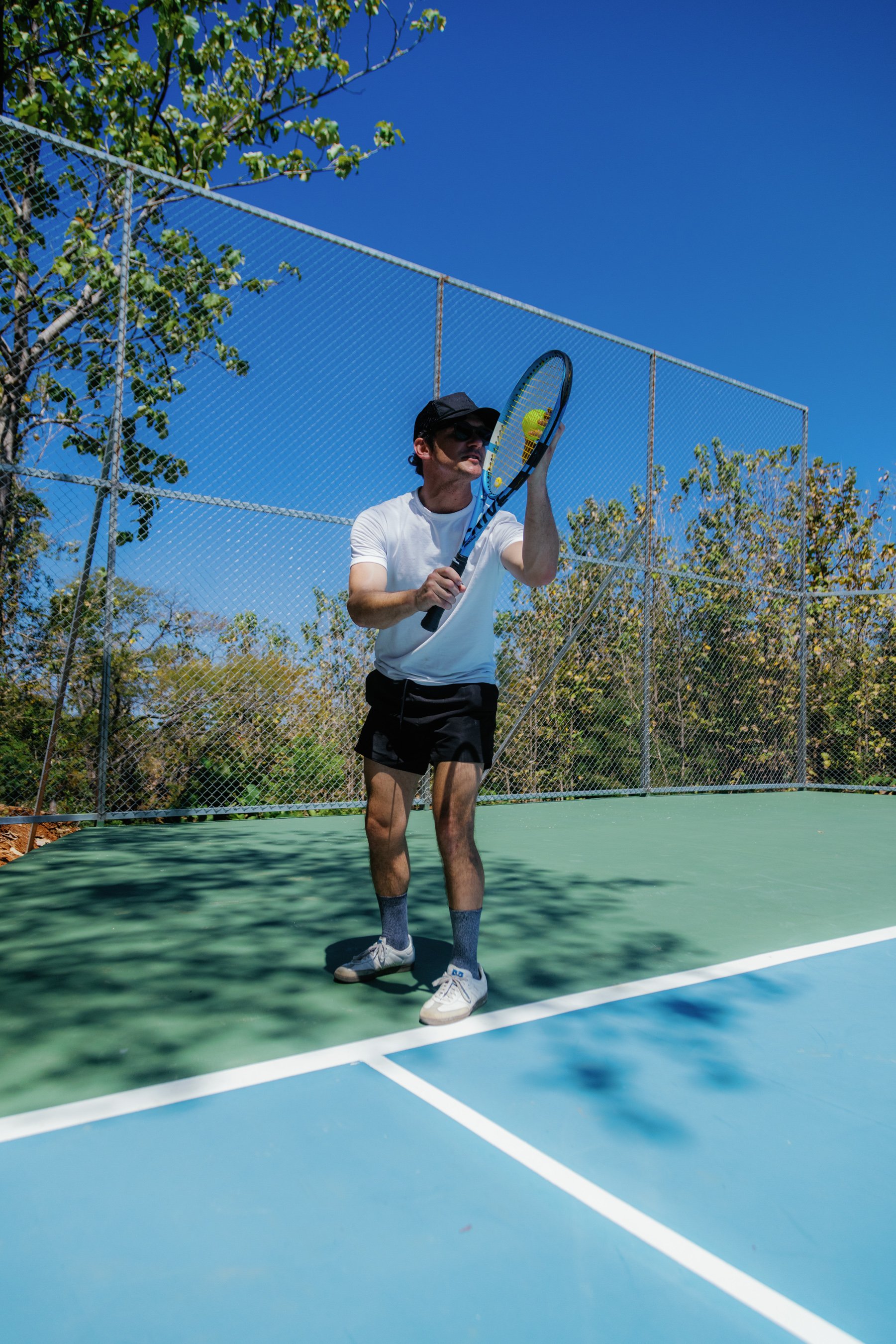 A man playing pickleball on a court, preparing to hit a yellow ball with a paddle under a clear blue sky.