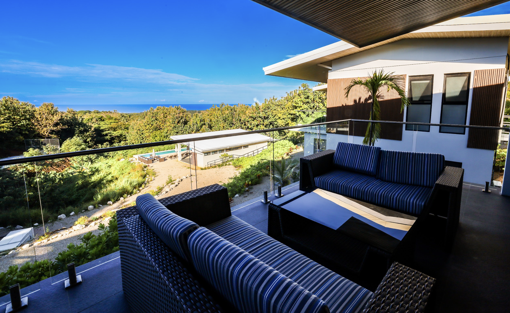 Balcony with outdoor furniture overlooking lush greenery and distant ocean on a sunny day.