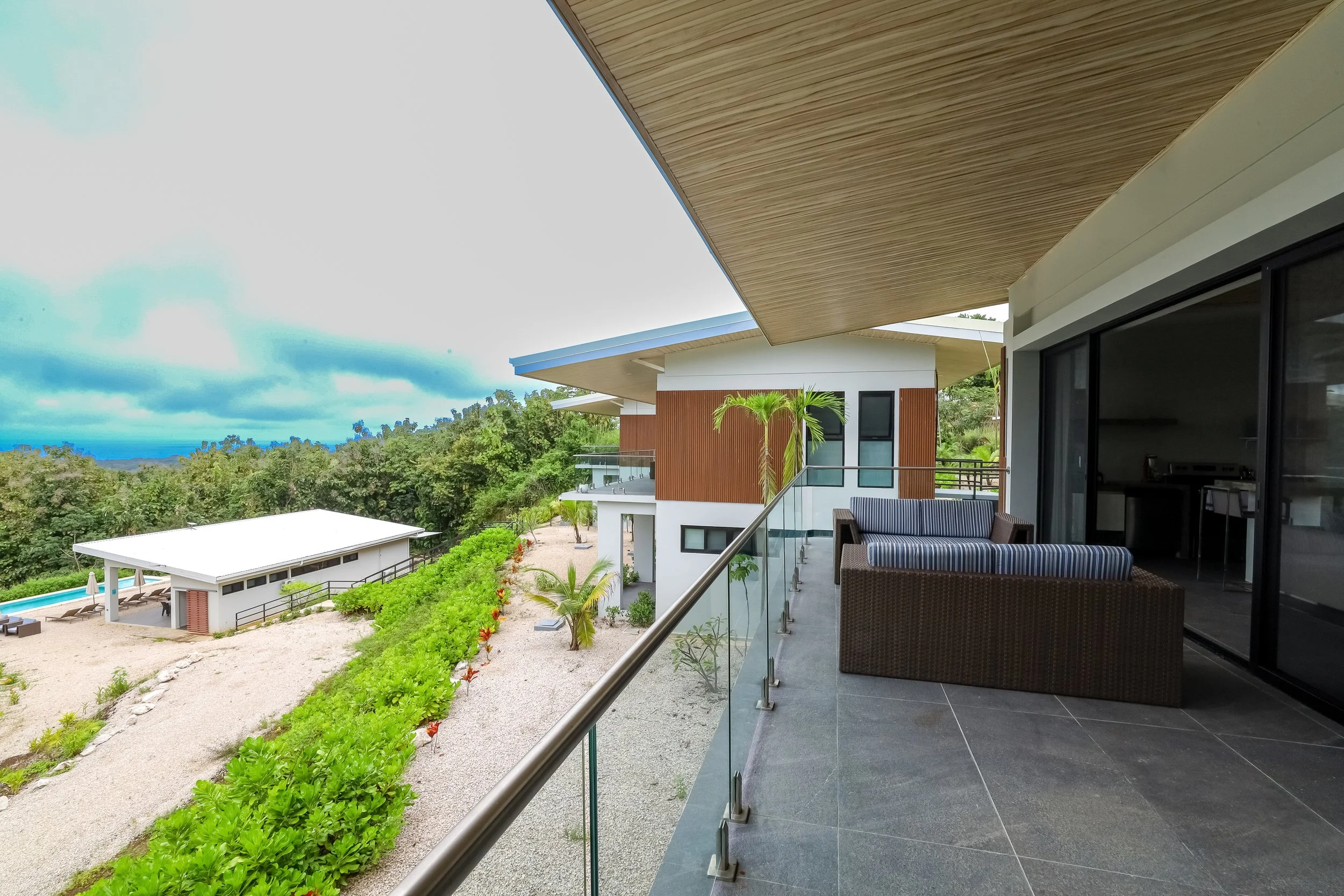Luxury villa balcony featuring a wicker outdoor sofa and glass railings overlooking lush greenery.