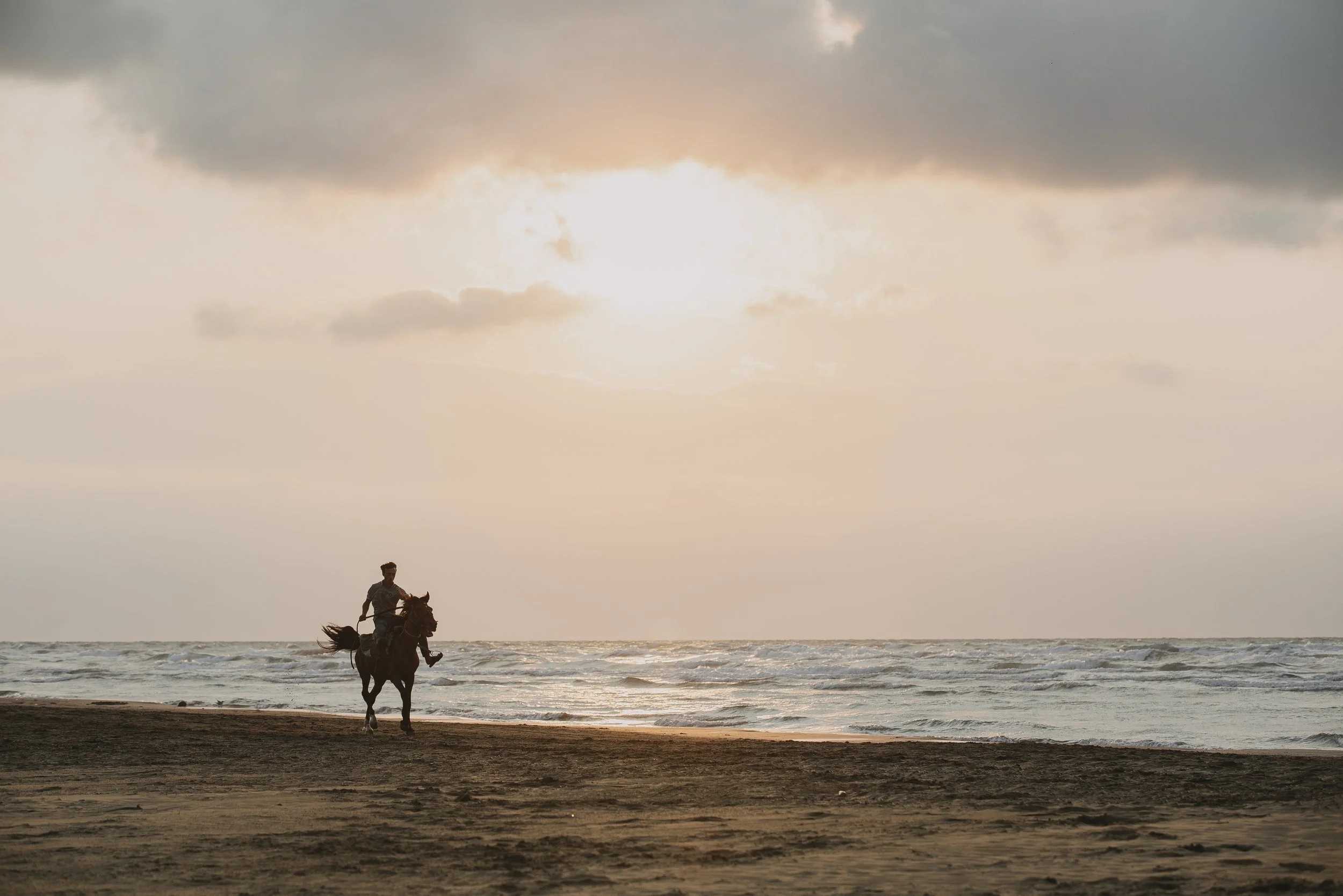 A person riding a horse along the beach at sunset, with the ocean waves in the background and a cloudy sky overhead.
