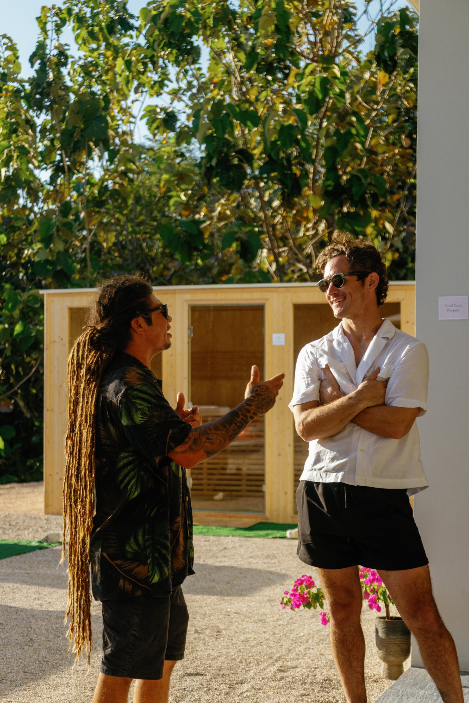 Two men engage in a friendly conversation outdoors, with one man pointing and the other laughing. One man has long dreadlocks and tattoos, wearing a black shirt with a green leaf pattern, while the other has short curly hair, wearing sunglasses, a wh