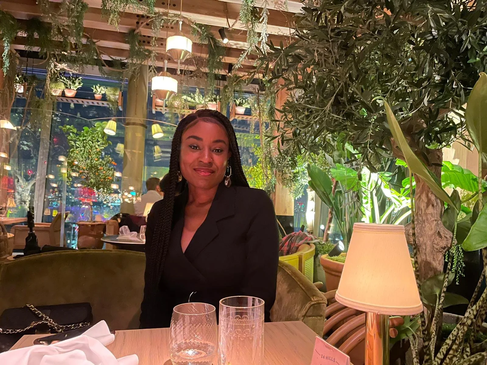 Woman sitting at a restaurant table with plants and hanging lights in the background.