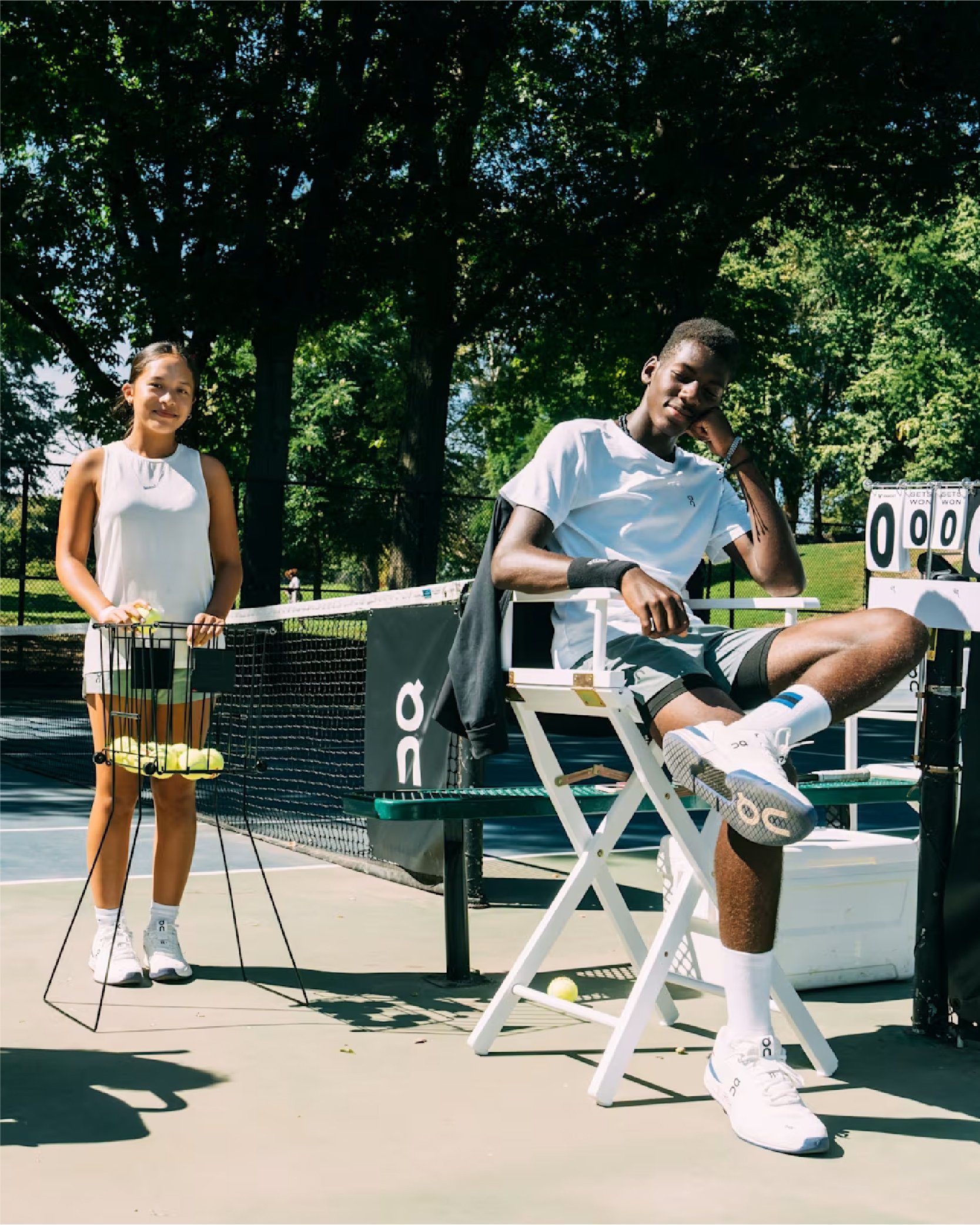 Two people on a tennis court; one is sitting on a chair with a foot up, and the other is standing, holding a tennis ball basket.