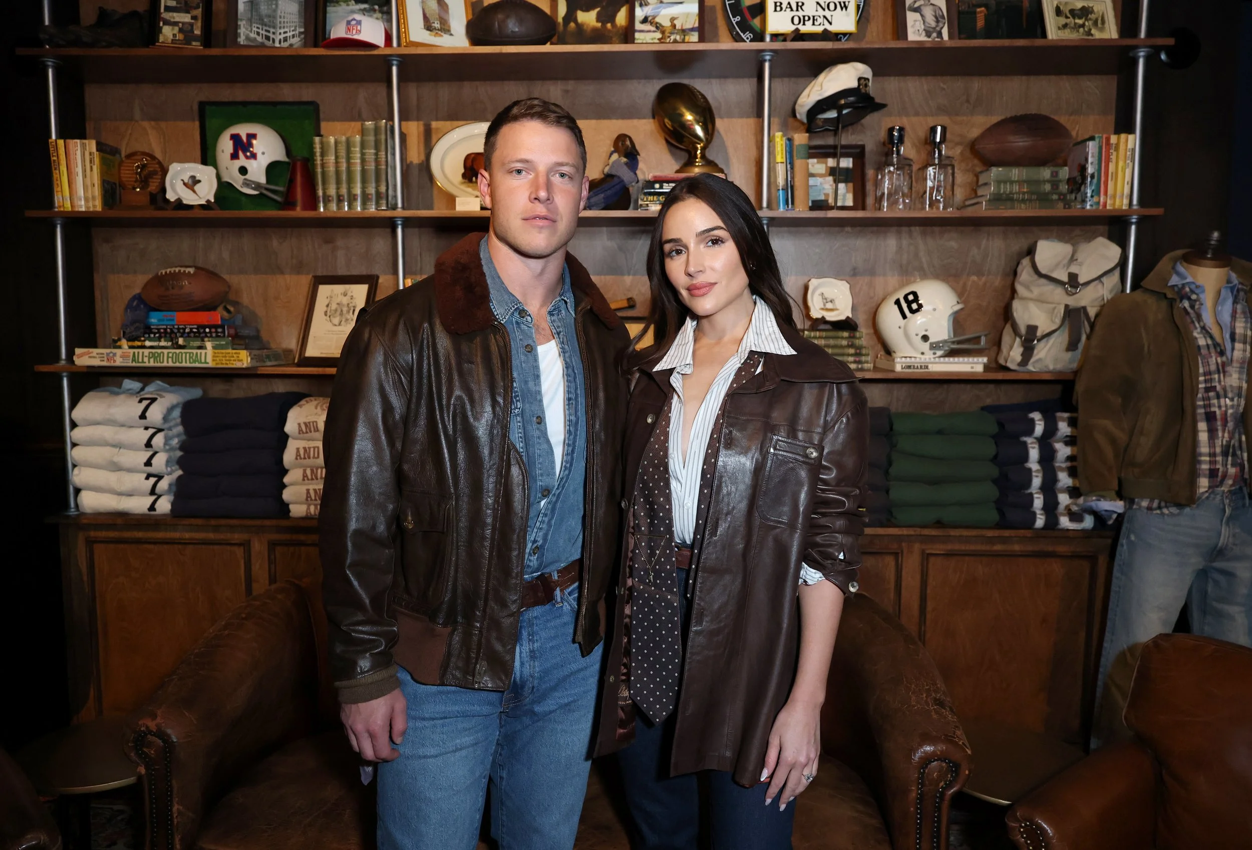 A man and a woman standing together in a room with shelves of books, football memorabilia, and vintage clothing in the background. They are both wearing leather jackets and posing for the photo.
