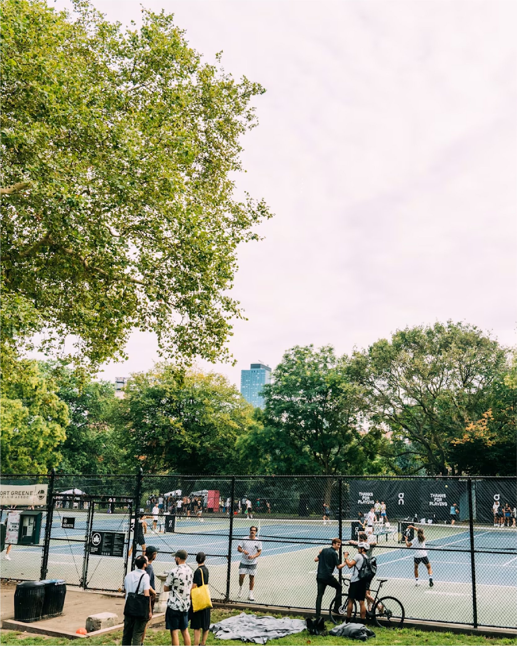 People watching a tennis match on outdoor courts surrounded by trees, with a city skyline visible in the background.