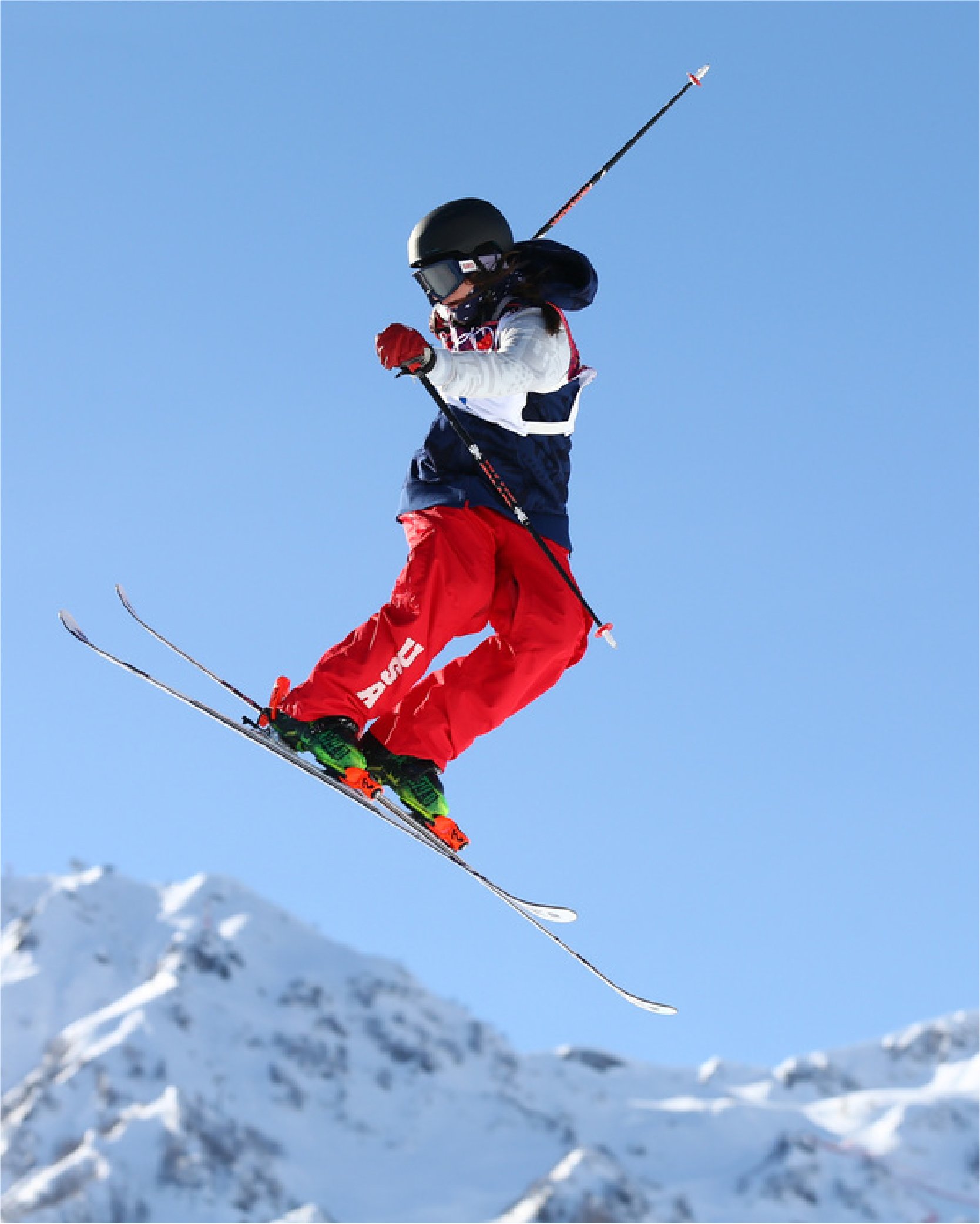Skier in mid-air performing a trick, wearing red pants and a helmet, against a backdrop of snow-covered mountains.