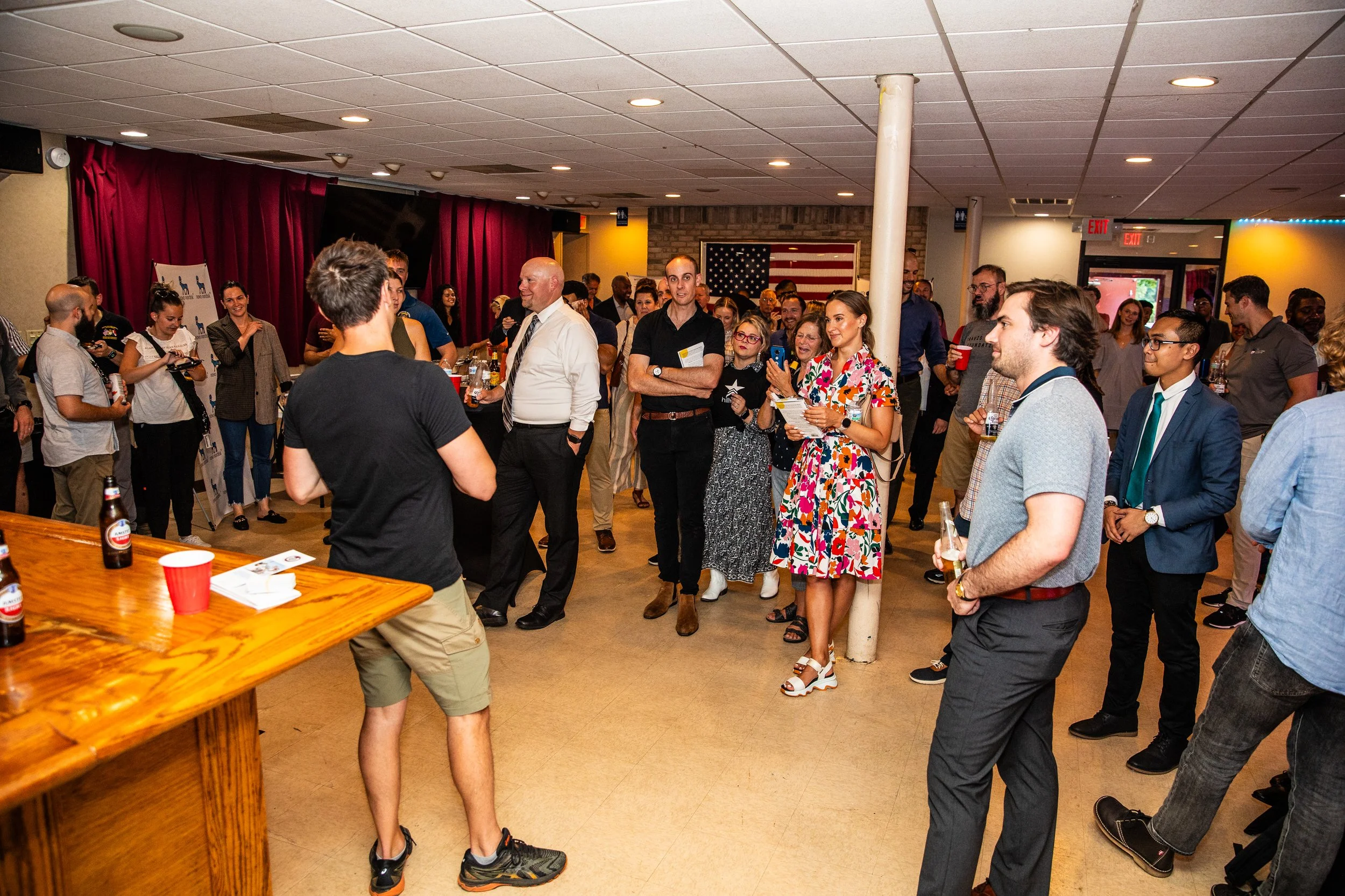 Group of people gathered at an indoor event, listening to a man speaking, with drinks and a wooden table in the foreground, red curtains, and American flag decoration in the background.