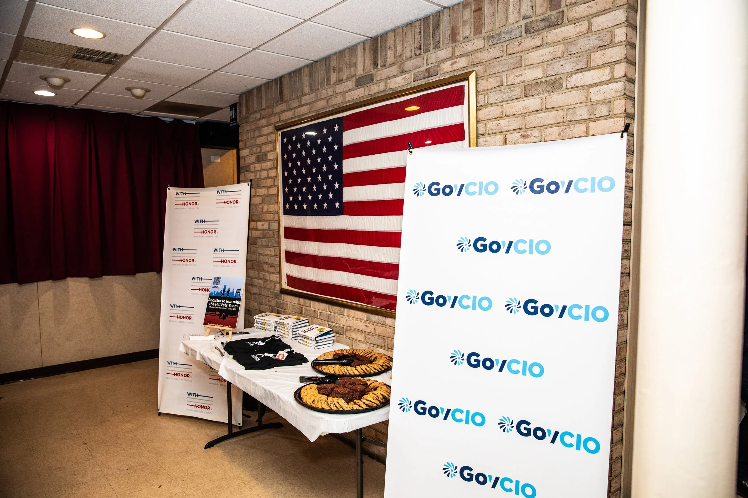 A table with snacks, brochures, and t-shirts in front of an American flag and two vertical banners with 'GovCIO' printed on them.