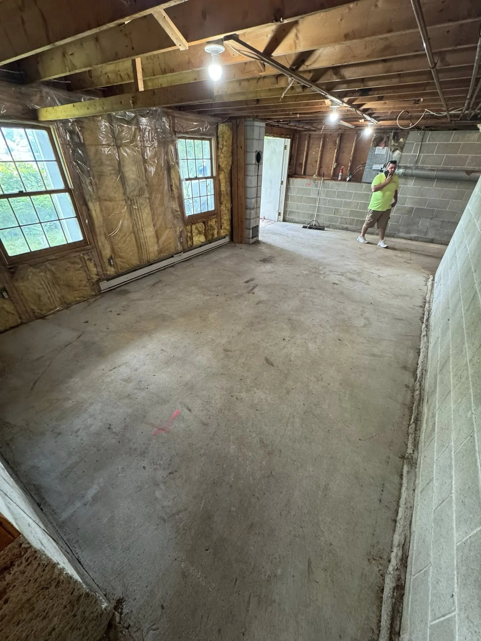 Interior of a house under renovation with exposed wooden ceiling beams, two windows, a concrete floor, and a person in a neon yellow shirt standing near a cinder block wall.