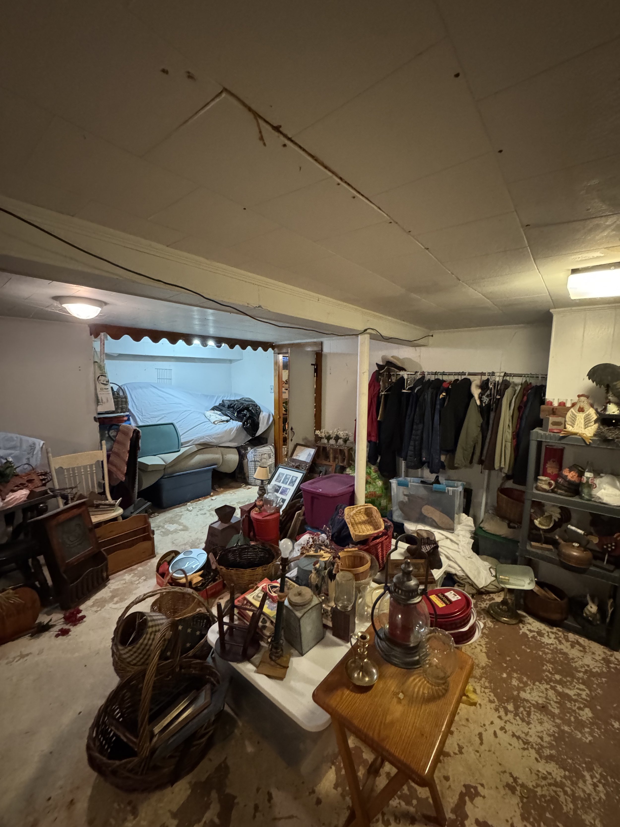 A cluttered basement room filled with various household items, furniture, and clothes, with a bed partly visible in the background and a low ceiling with a crack.