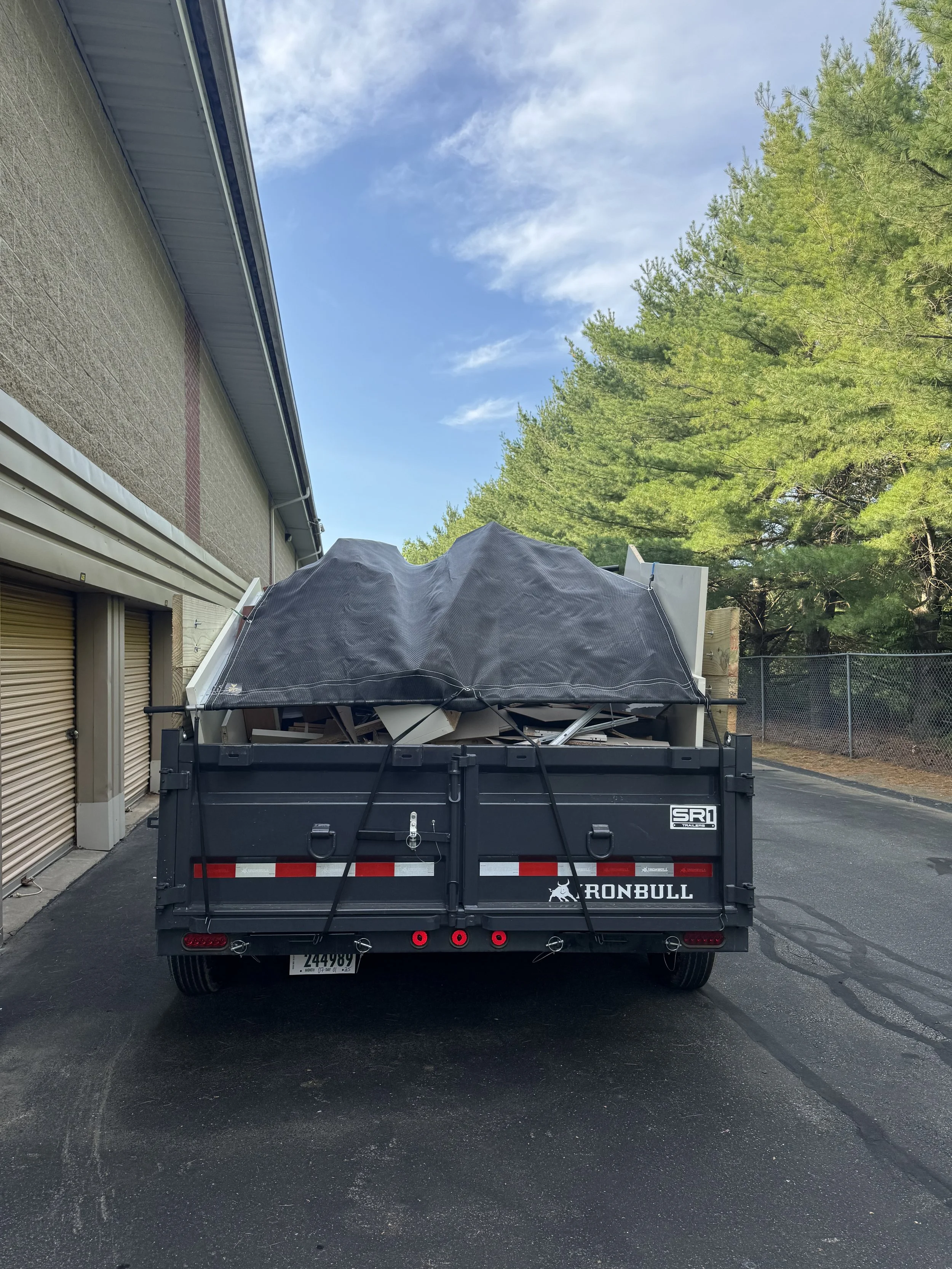 The back of a black Iron Bull utility trailer filled with various debris, including a large black tarp, against a building wall with trees and a blue sky with clouds in the background.