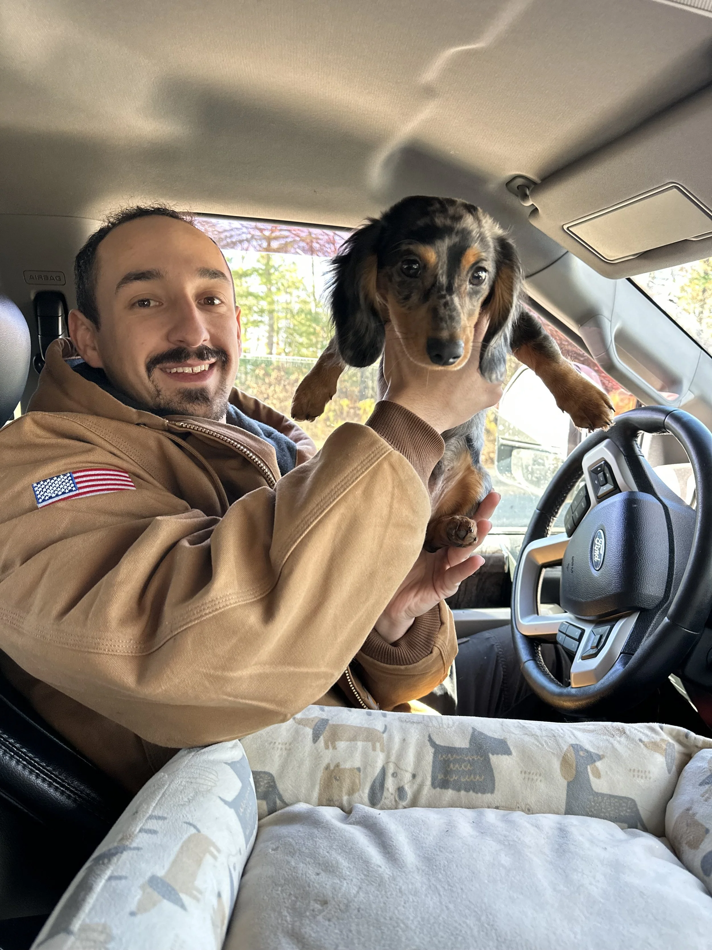 A man smiling and holding a dachshund puppy inside a vehicle with a beige interior and trees visible outside the window.