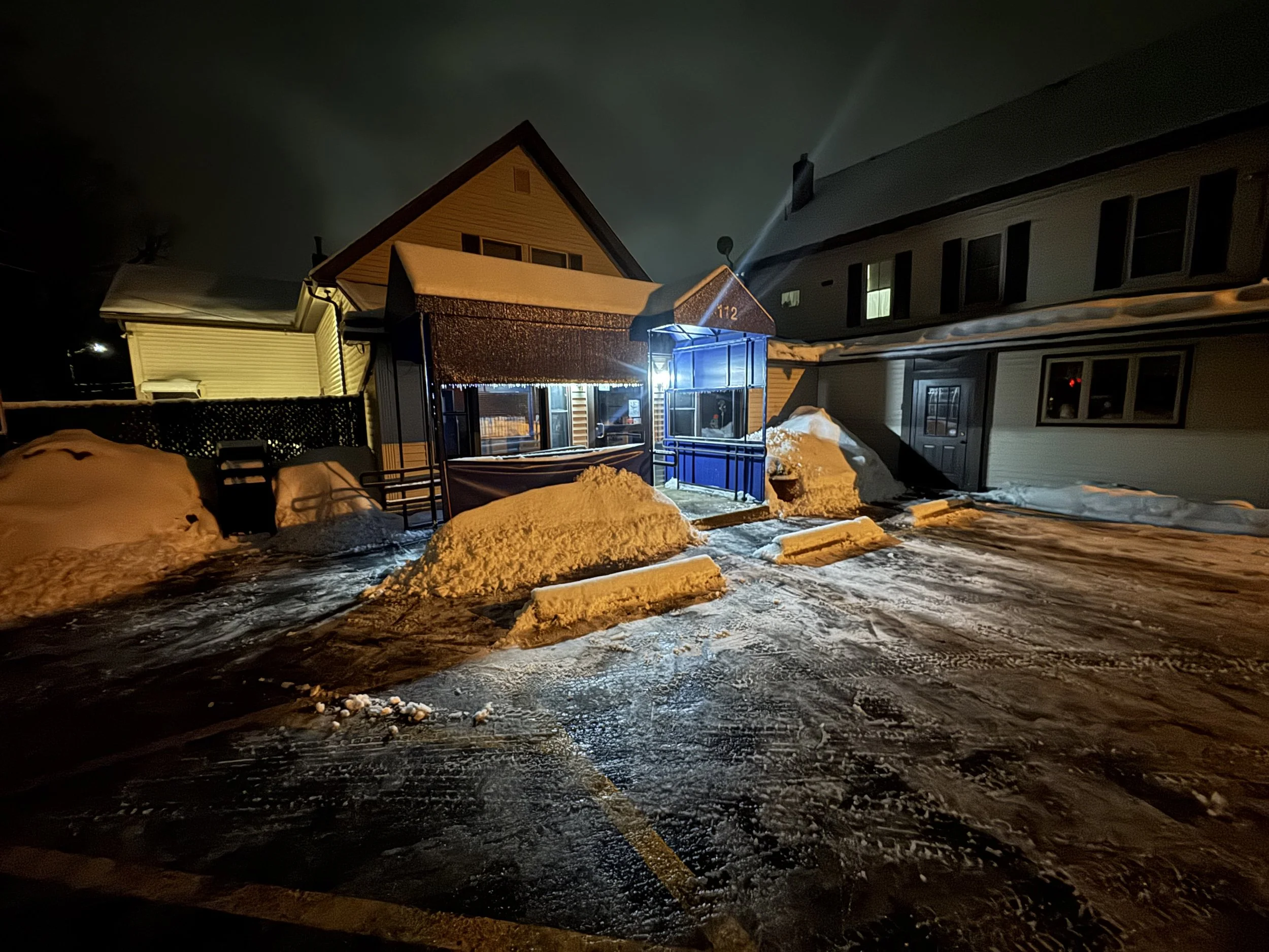 Snow-covered parking lot at night, with a building featuring a lit entrance and a drive-thru window, surrounded by snow piles and tire tracks.
