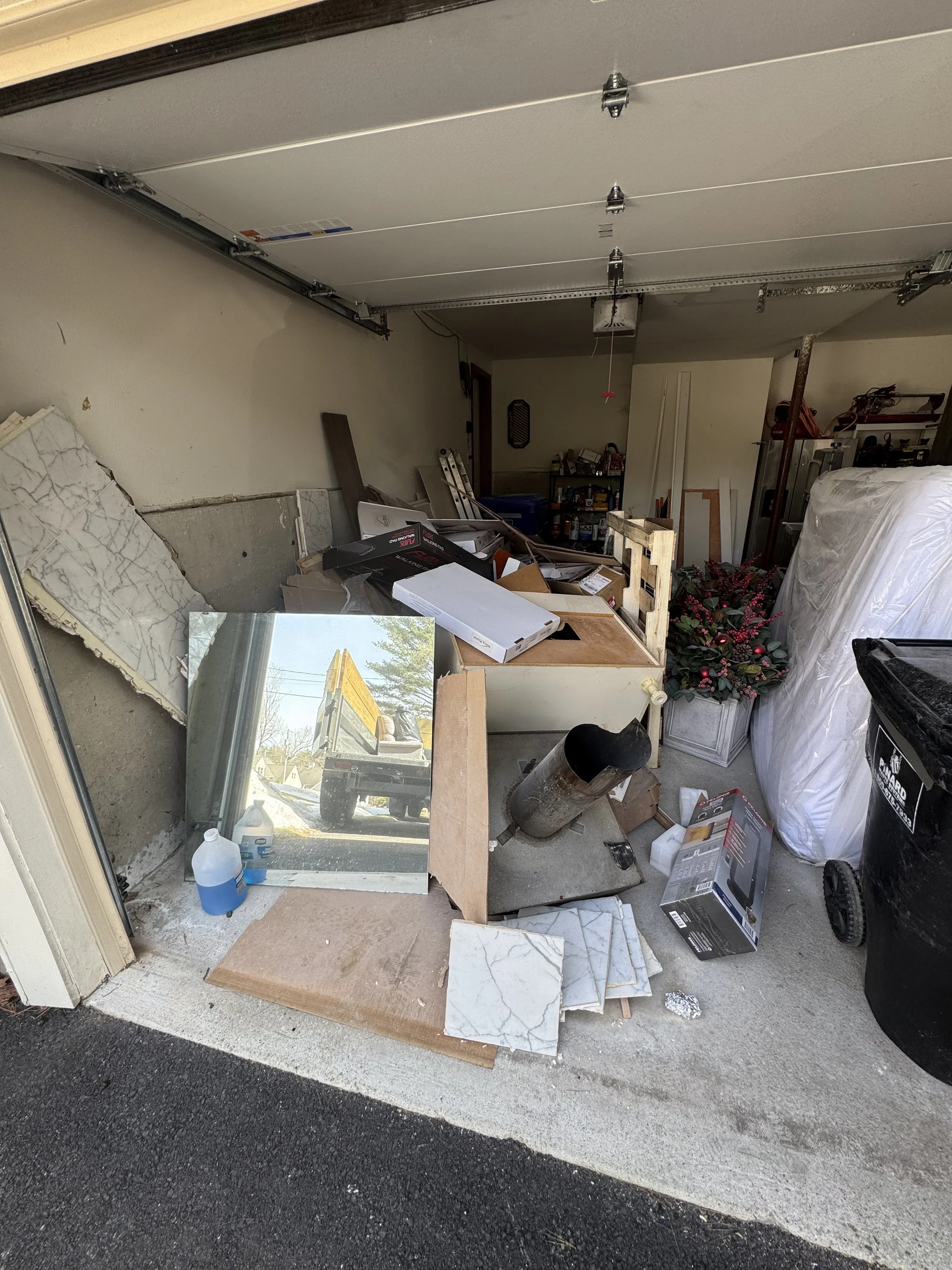 Garage with clutter, including tiles, a mirror, various boxes, a potted plant, a black trash bin, and construction materials.