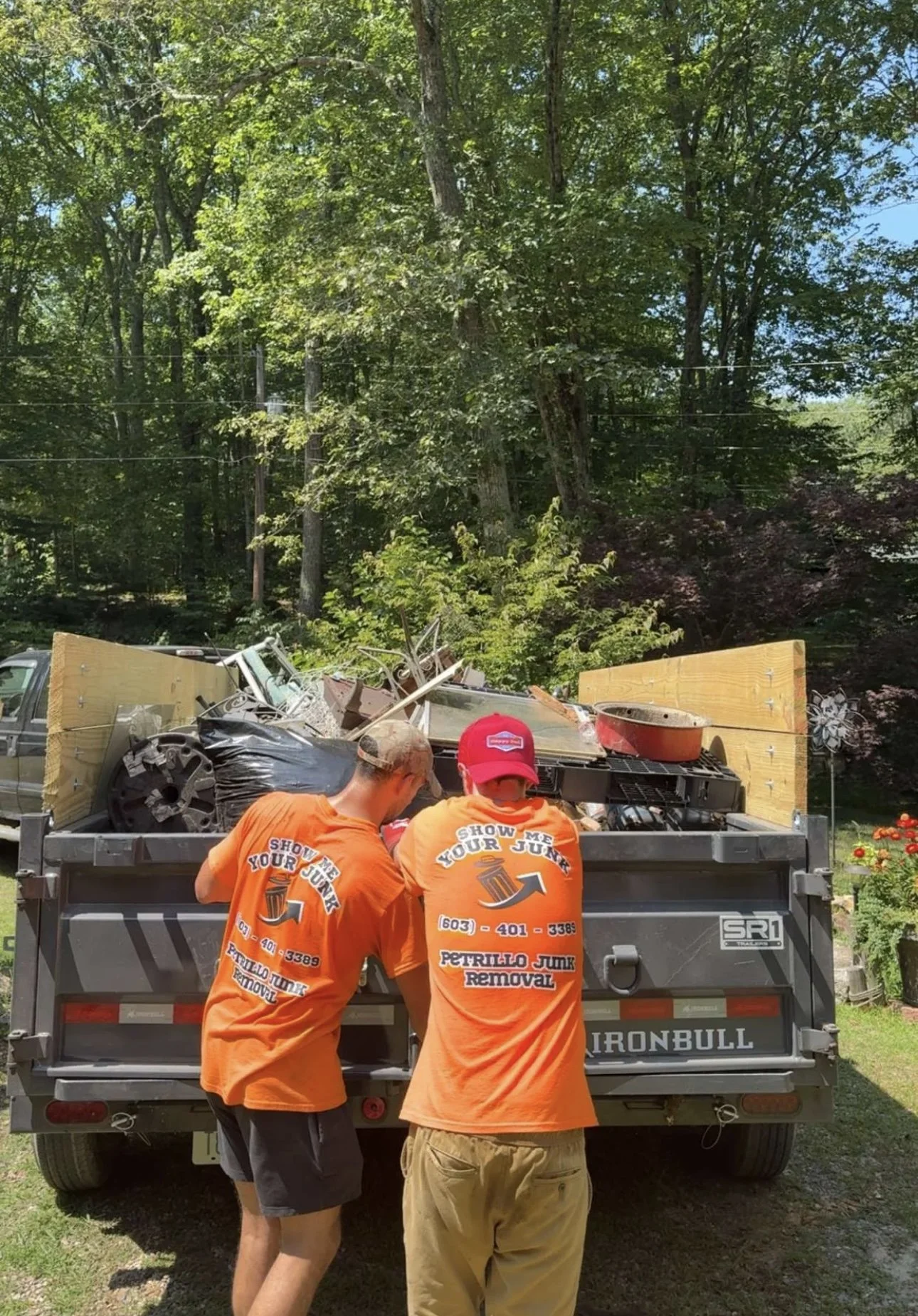 Two workers from Petrillo Junk Removal wearing orange shirts loading a truck with scrap metal and debris. The trucks has Wood paneling on the sides and the motto "Show Me Your Junk" along with contact information.
