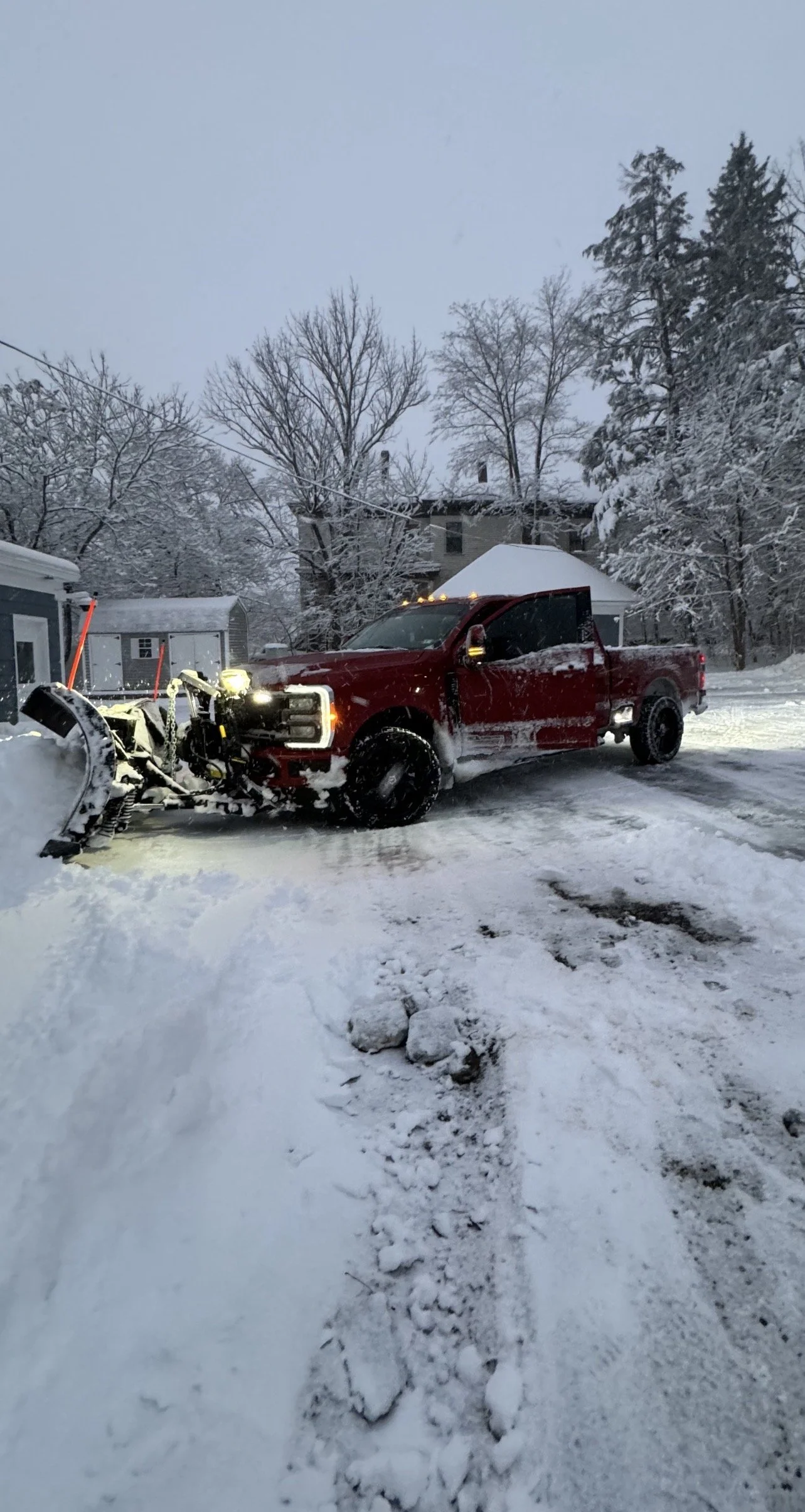 A red pickup truck parked in a snowy driveway, with snow removal equipment attached to the front. Snow-covered trees and a house are visible in the background during winter.