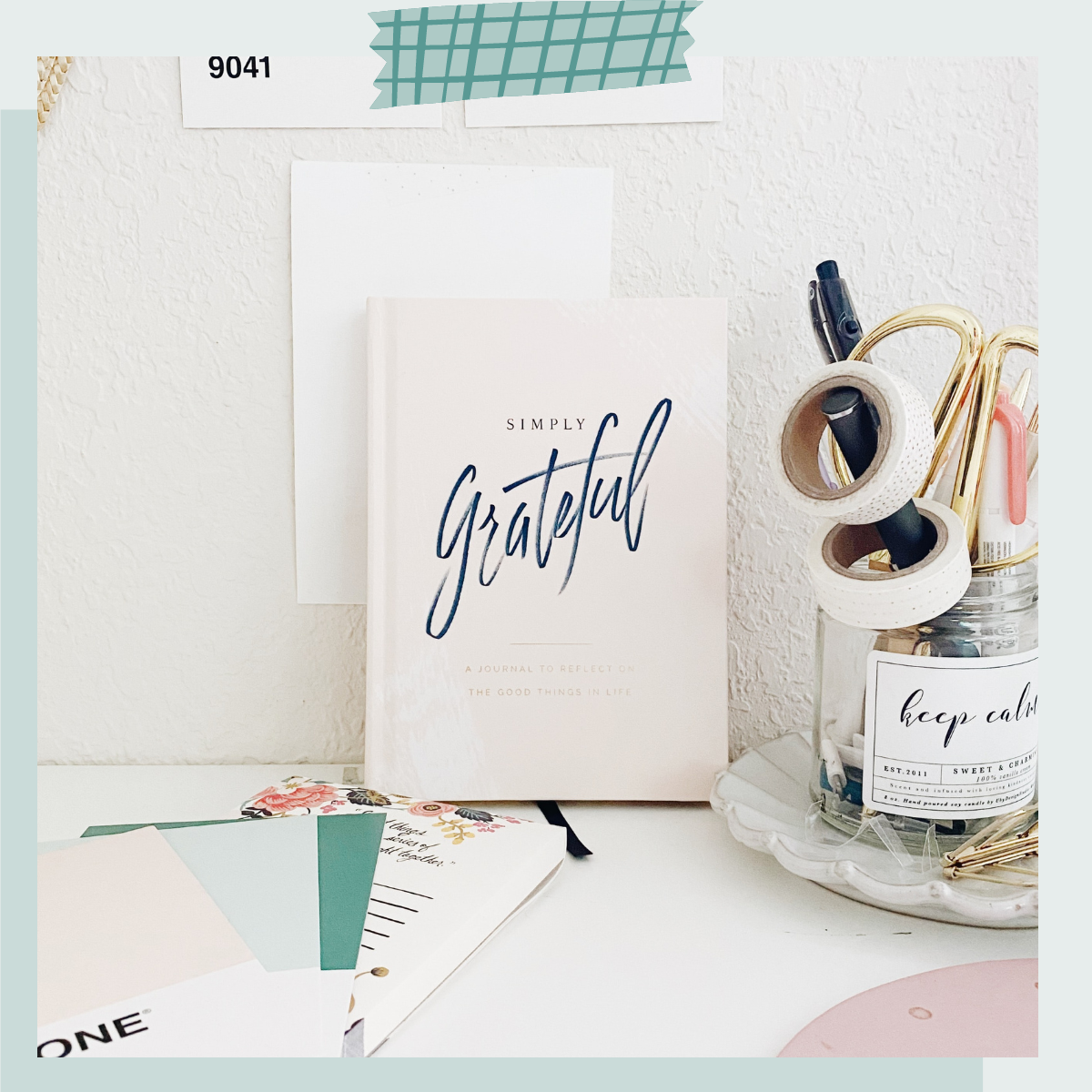 A desk with a notebook titled 'Simply Grateful,' a jar labeled 'keep calm' filled with pens and paper clips, and colorful stationery papers.