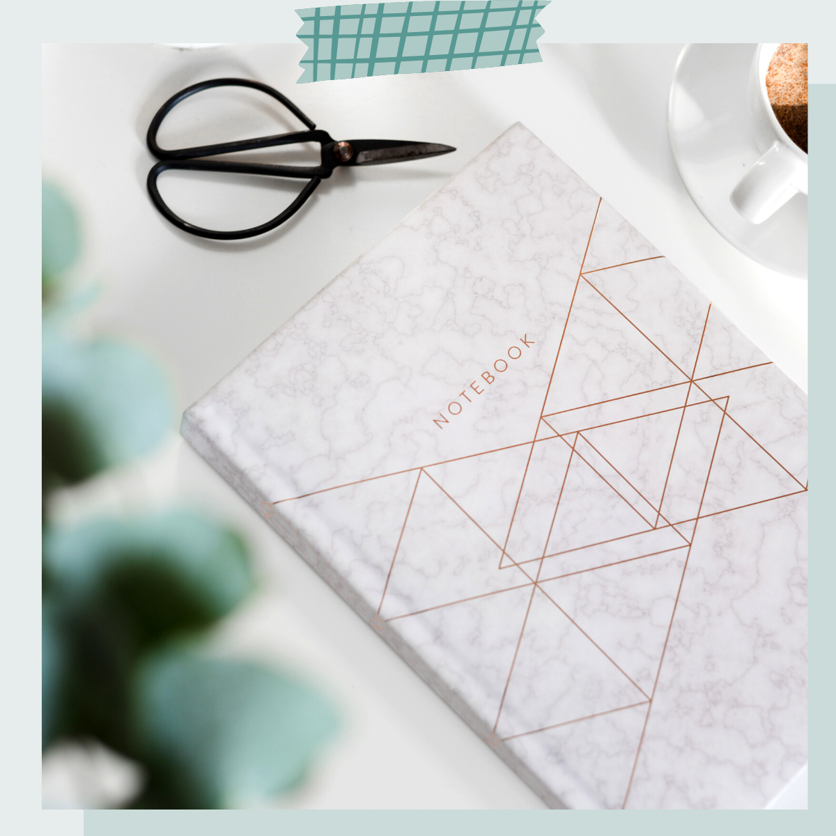 A marble-patterned notebook with geometric lines on the cover, a pair of black scissors, a cup of coffee, and a piece of green grid tape on a white surface.