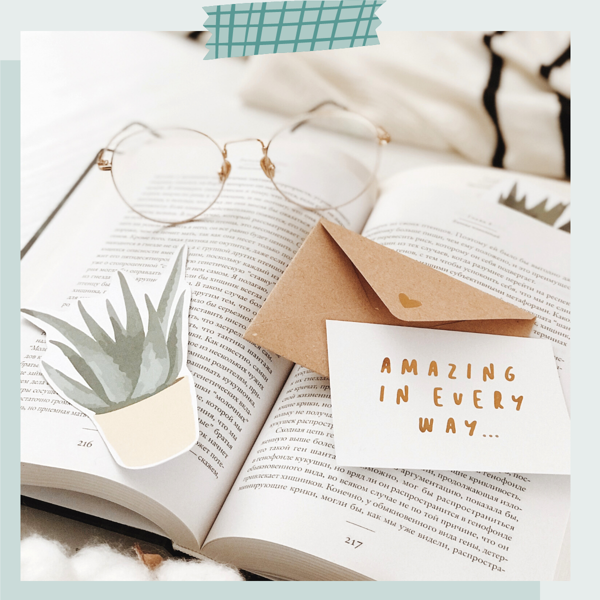 An open book with reading glasses, a potted plant illustration, a small envelope, and a card with the message "AMAZING IN EVERY WAY" on a soft background.