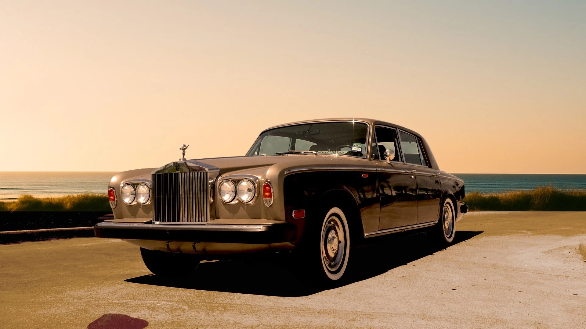 A vintage black and beige luxury car parked on a coastal road with the ocean and a clear sky in the background.