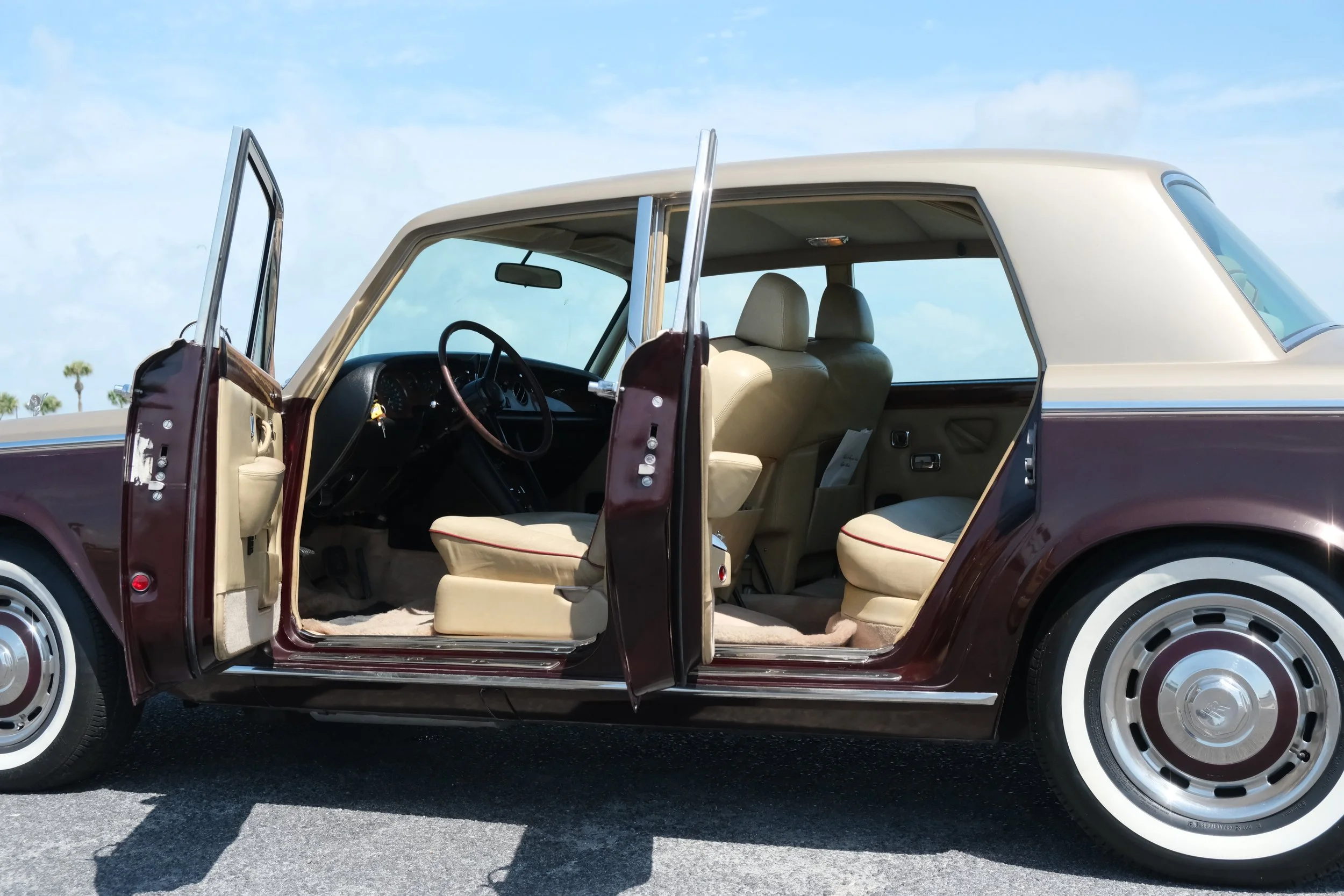 A vintage two-tone beige and maroon station wagon with all four doors open, showing beige interior seats and dashboard, parked on a paved surface under a cloudy sky.