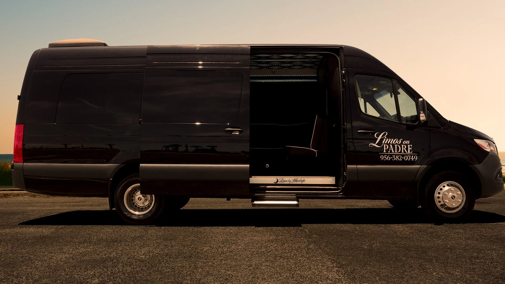 Black delivery van with sliding side door open, advertising "Limos on PADRE" and a phone number, parked on a paved road with a clear sky in the background.